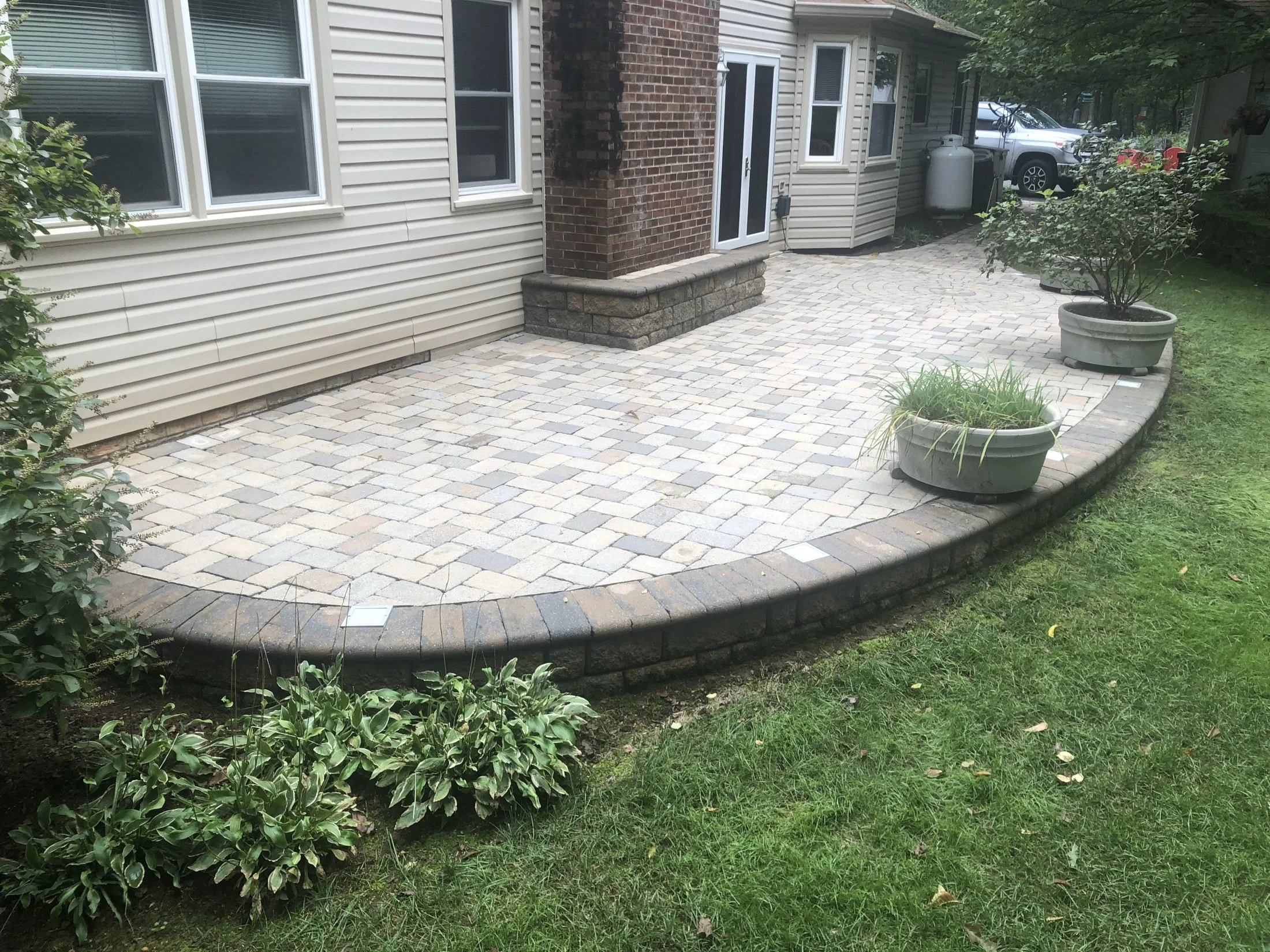 Raised backyard patio with multicolored pavers, brick edging, house siding, potted plants, and surrounding lawn