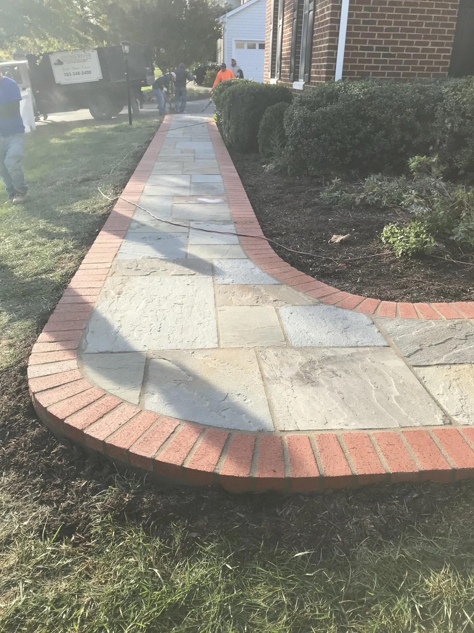 Landscaping crew installing a curved brick-edged flagstone walkway beside a brick house.