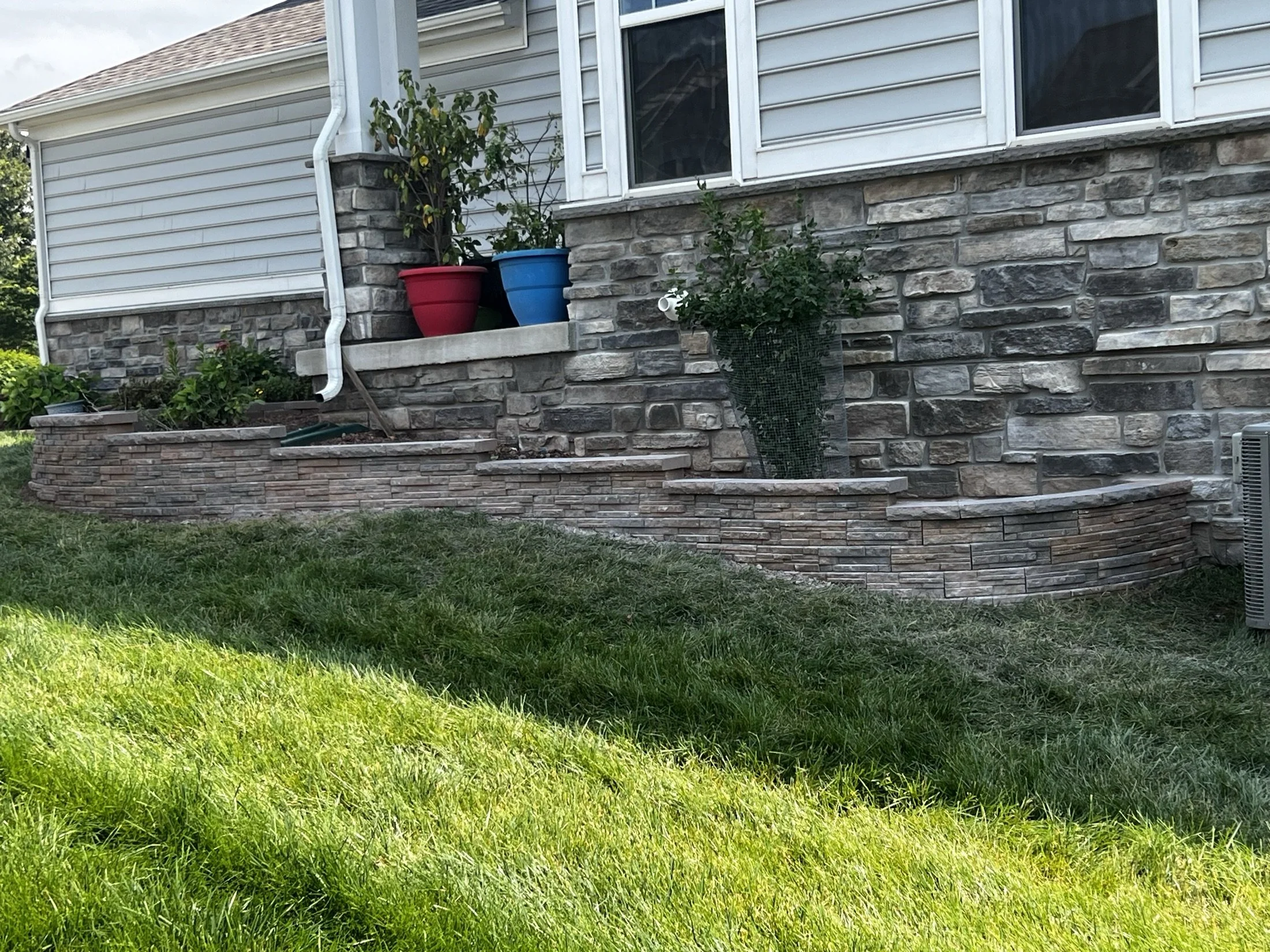 Stone-faced house foundation with raised garden beds, potted plants, and a grassy lawn