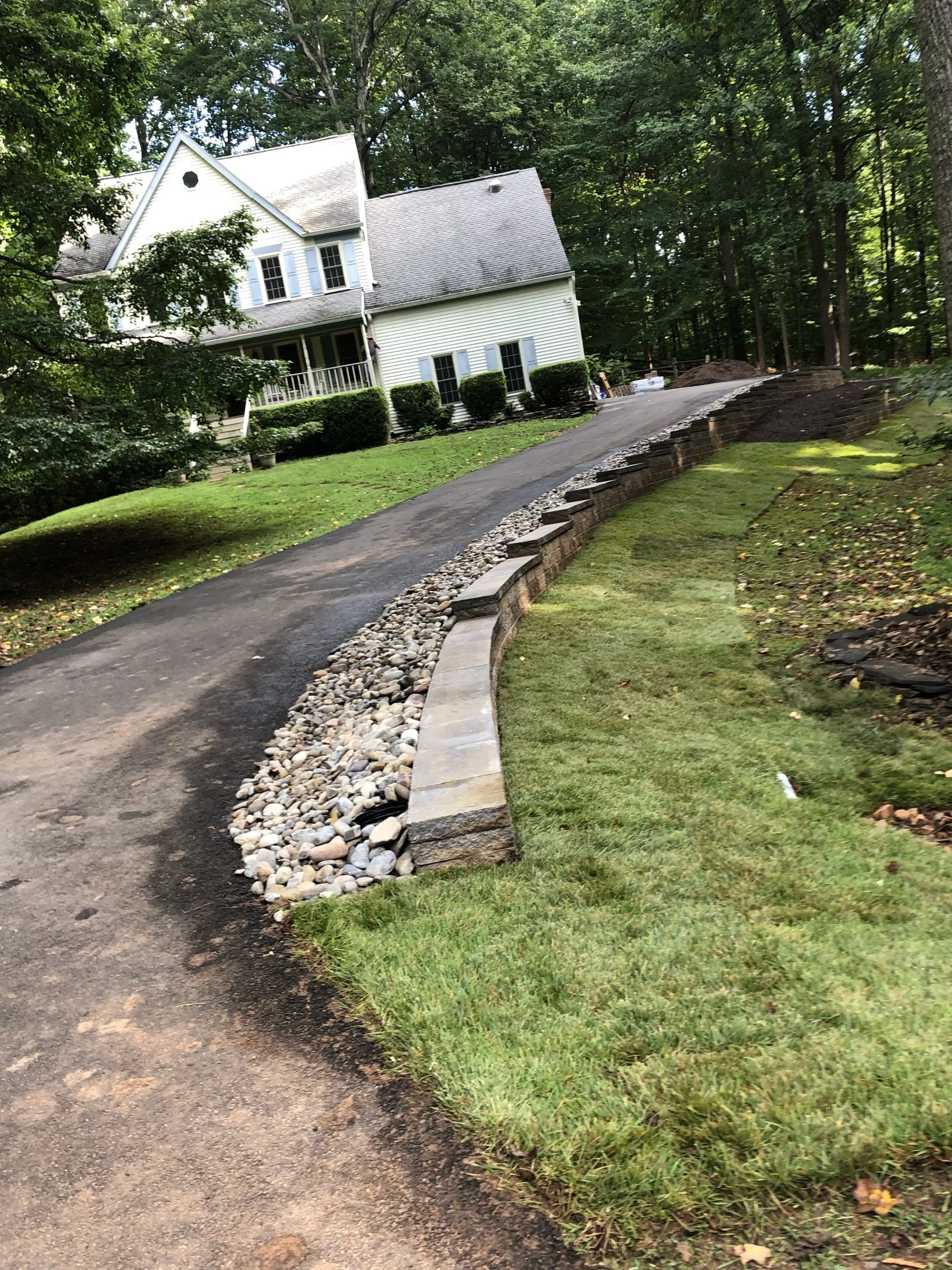 White suburban house with a front porch, paved driveway, manicured lawn, stone retaining wall, and wooded surroundings