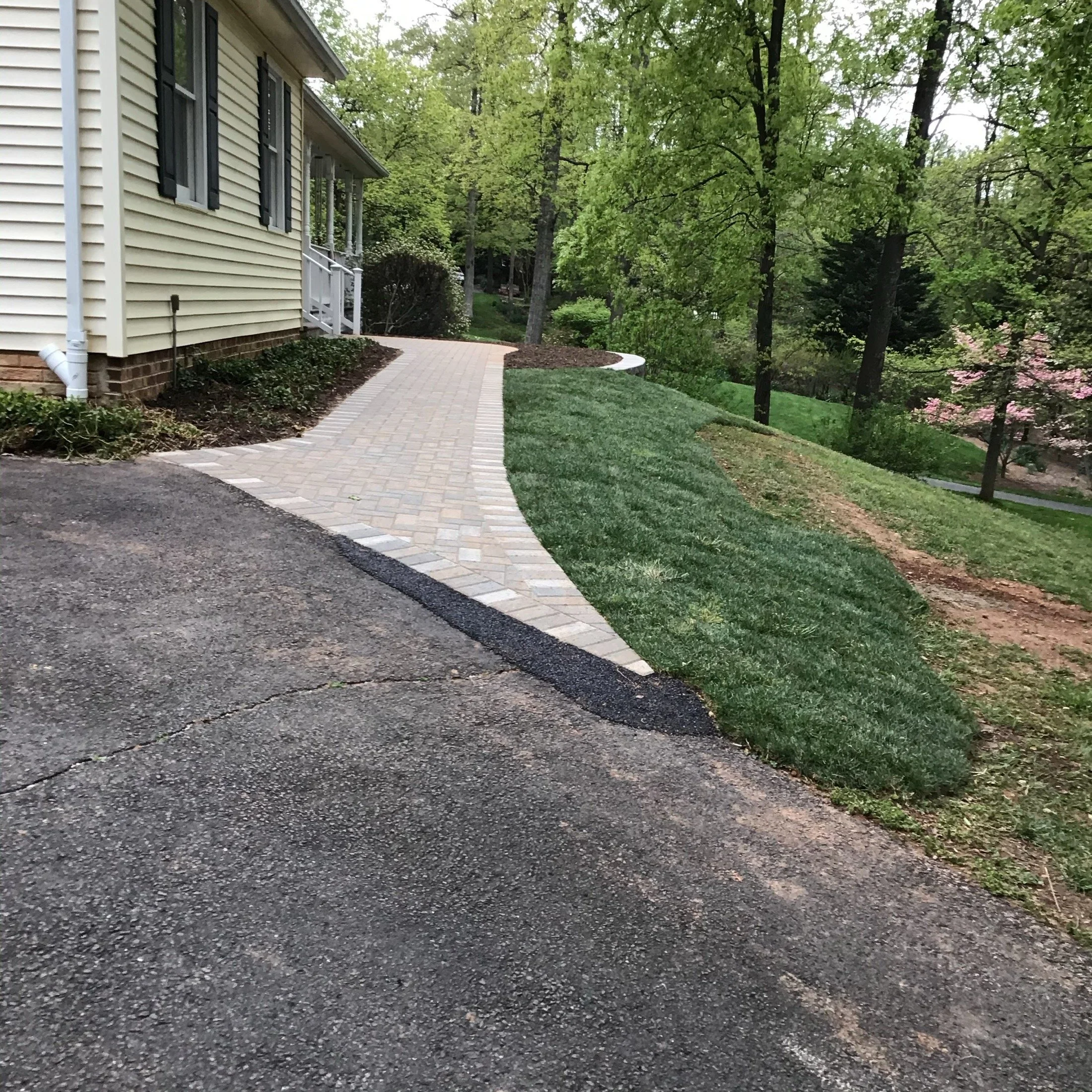 Brick walkway beside a beige house leading through a landscaped, tree-filled yard