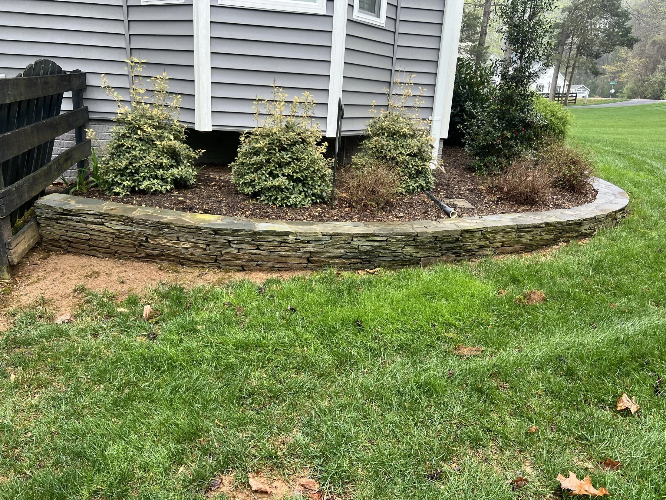 Landscaped garden bed with shrubs beside a gray-sided house, bordered by a curved stacked-stone retaining wall and green lawn.