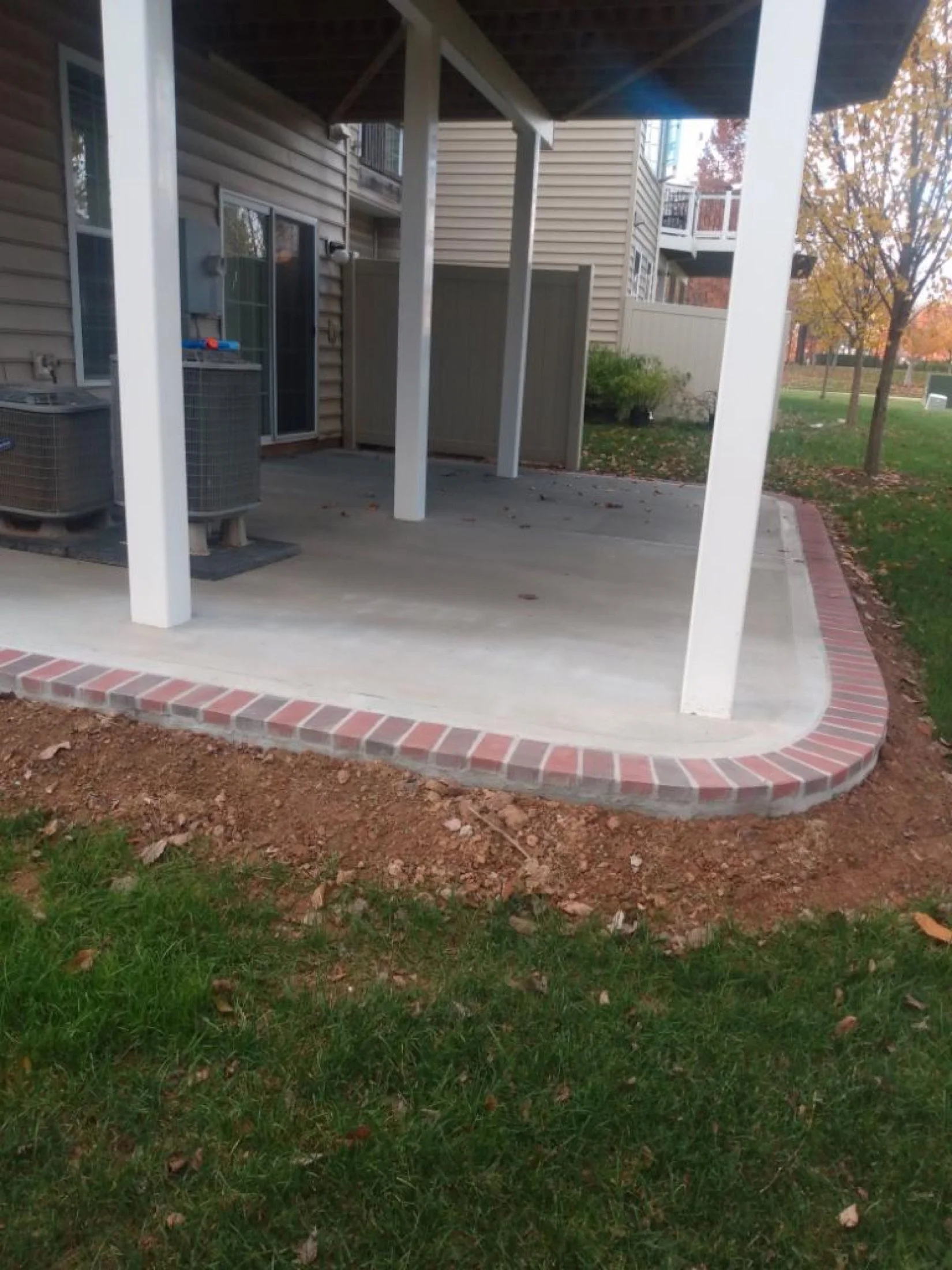 Covered concrete patio with white support posts, brick edging, outdoor air-conditioning units, and a townhouse exterior.
