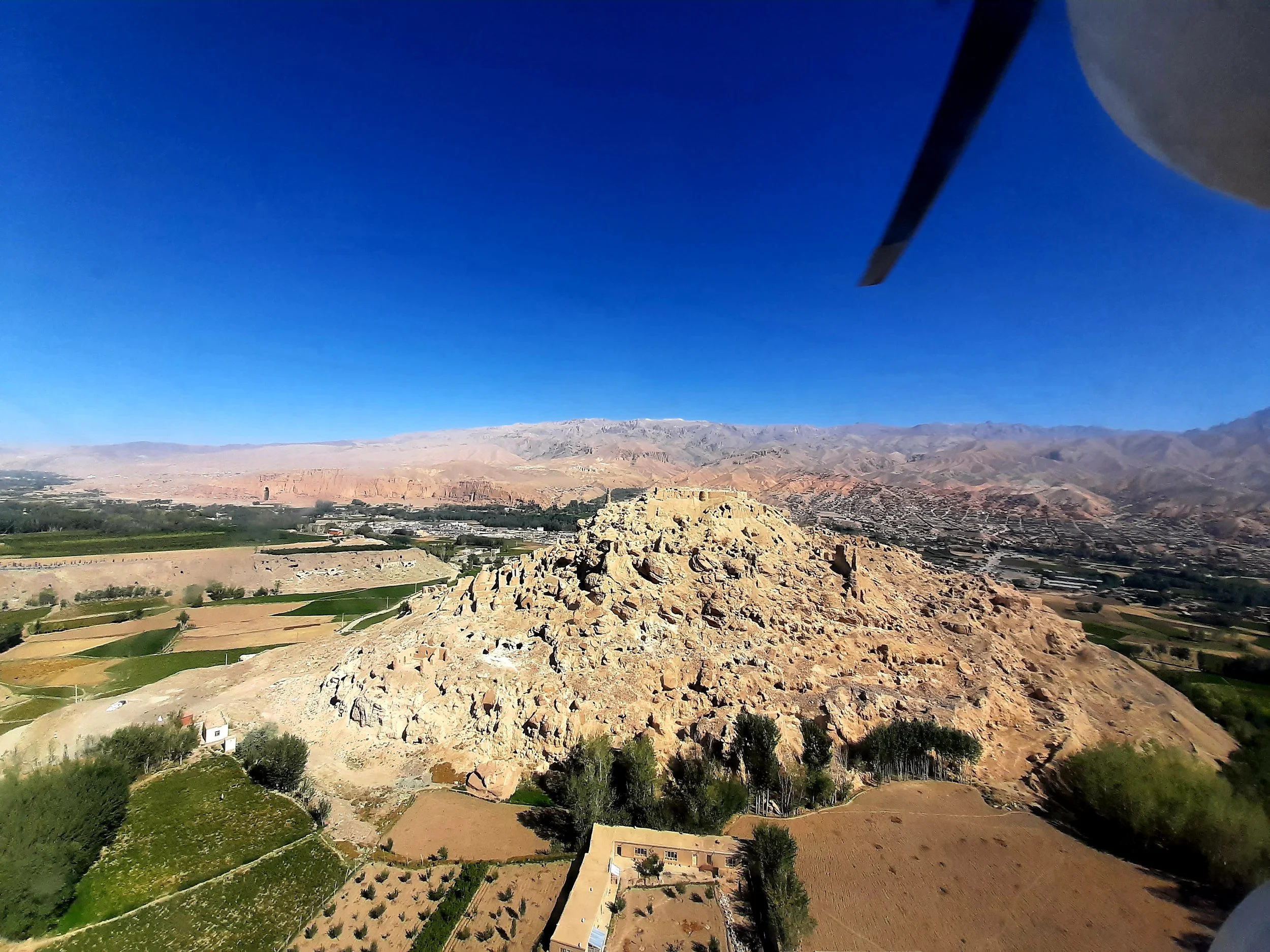 An aerial view of Shahr-e-Gholghola (Gholghola City), the historic hilltop ruins in the foreground, overlooking Bamyan’s rugged landscape, fertile fields and surrounding valley.
