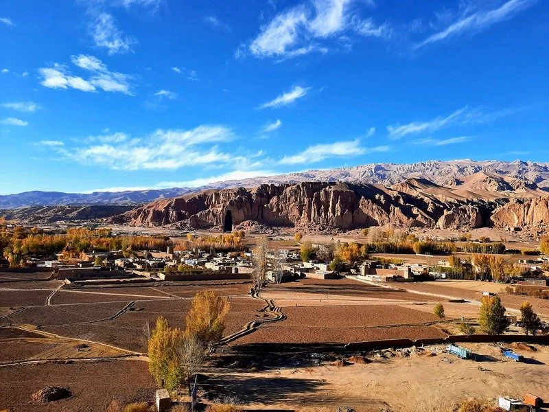 A view across the Bamyan valley from the Bamyan Cultural Center (BCC) area, with cultivated fields and traditional settlements framed by the dramatic sandstone cliffs and the monumental niches of the Buddhas of Bamyan. The landscape reflects the regi