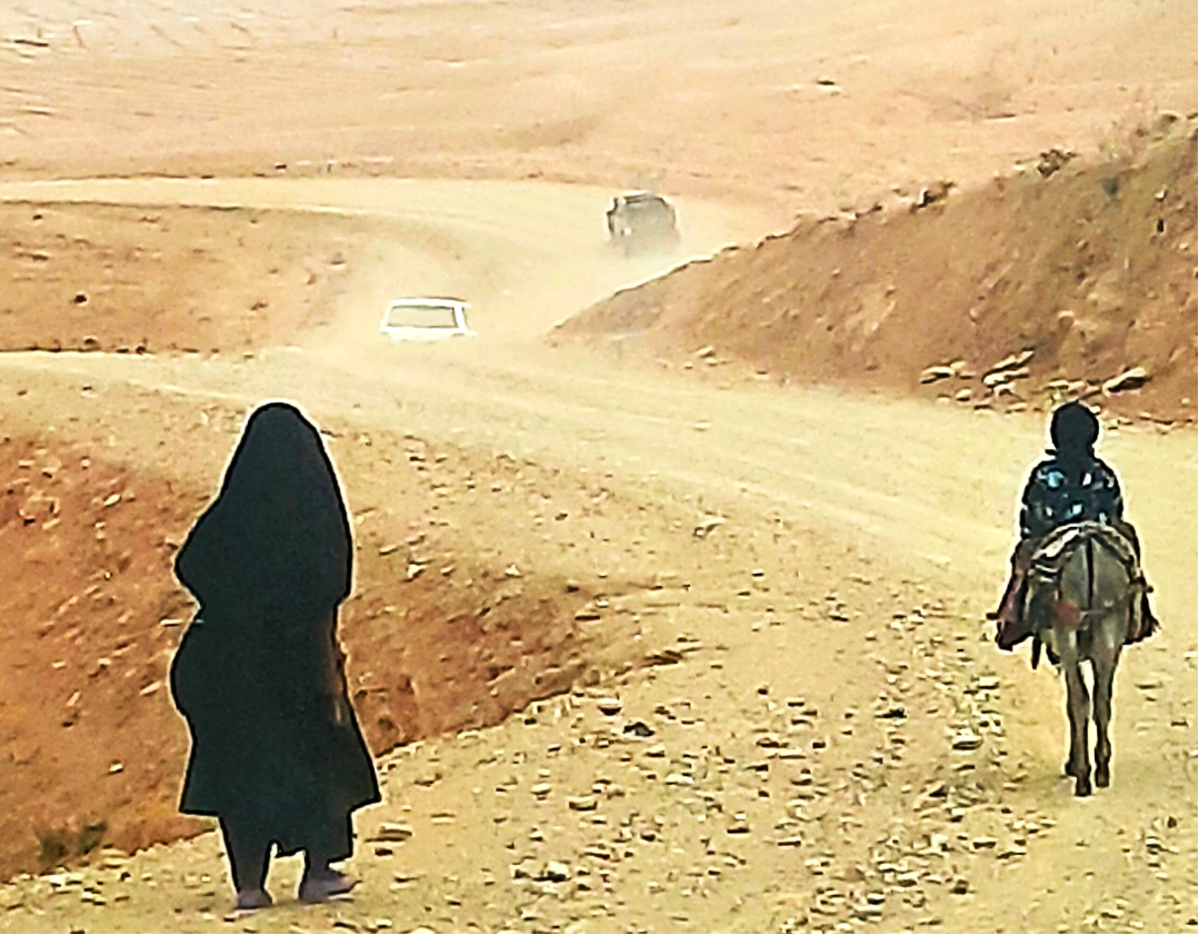 A woman walks along a dusty, sandy road while a child rides a donkey through the dry landscape of Daikundi. In this mountainous region, where distances are long and roads can be difficult, walking and donkeys remain part of everyday life, connecting 