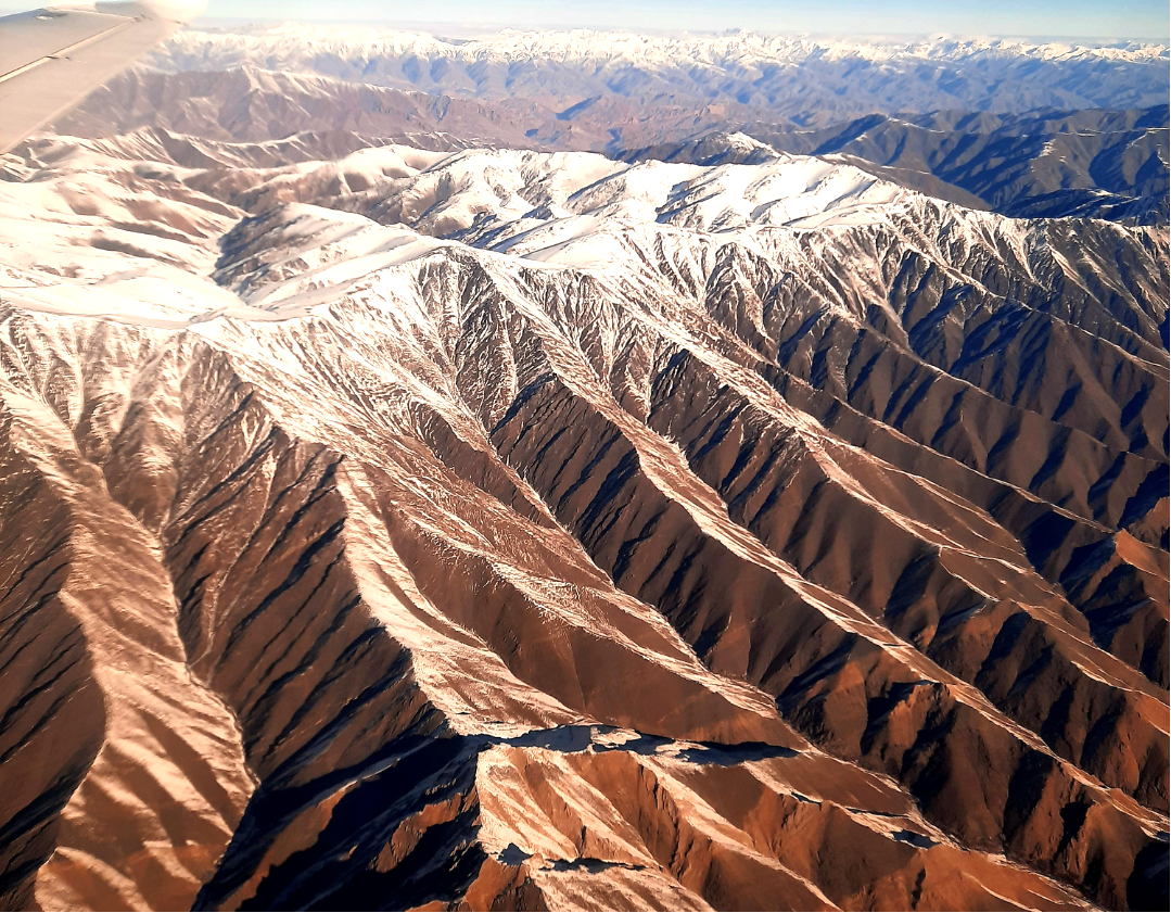 Successive mountain ridges stretch towards the horizon, their steep slopes carved into intricate lines of ochre, rust and shadow. The aerial perspective reveals the immense scale and geological character of this remote landscape, where the mountains 