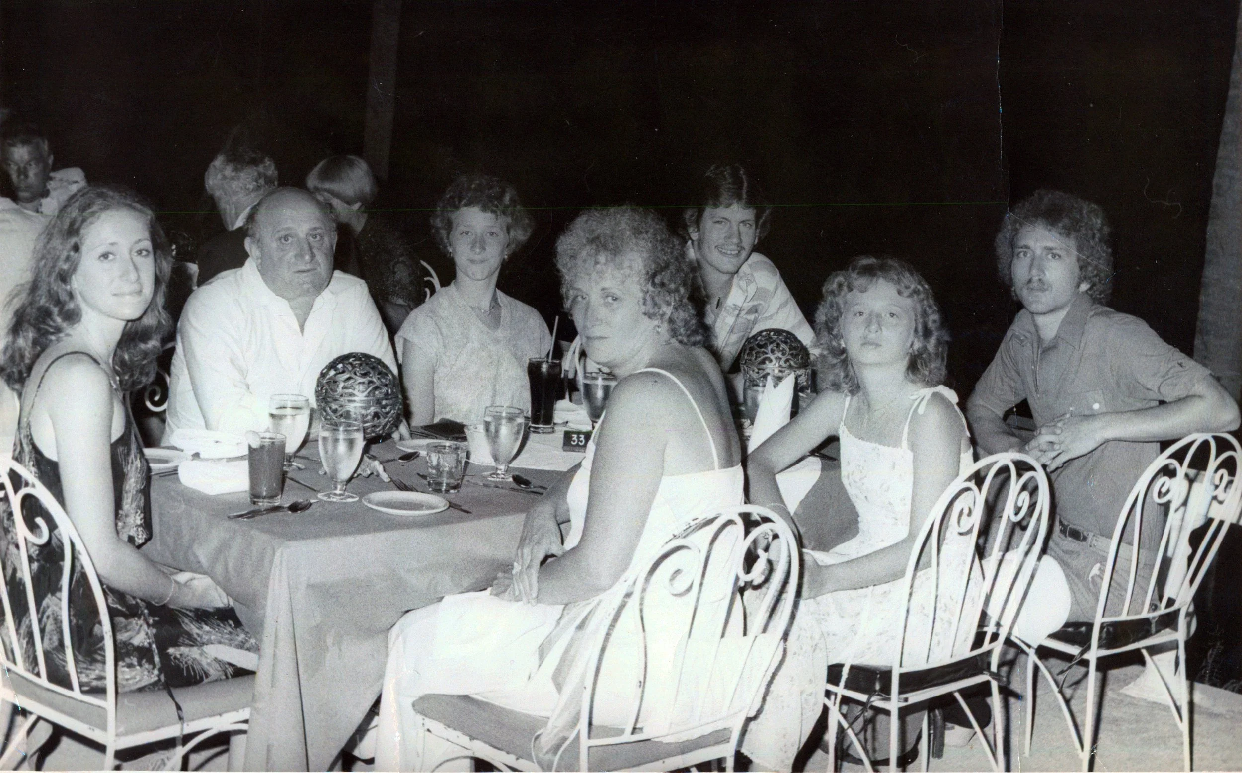 A black-and-white photo of people sitting at a dinner table outdoors, under trees, during nighttime. The table has drinks and tableware, with a group of women and men looking at the camera.