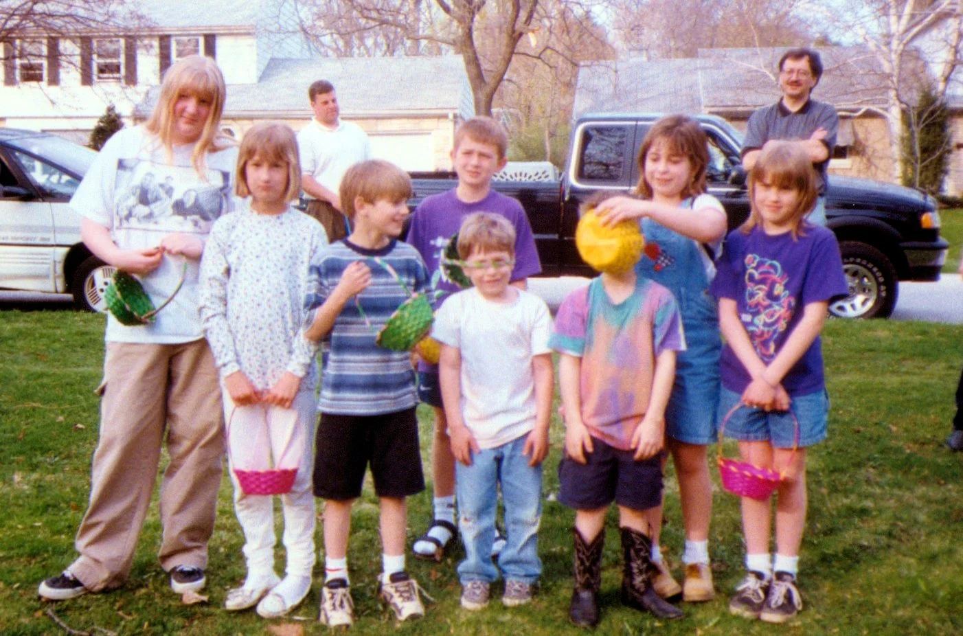 Group of children and two adults outdoors on a grassy area, holding Easter baskets and eggs during daytime.