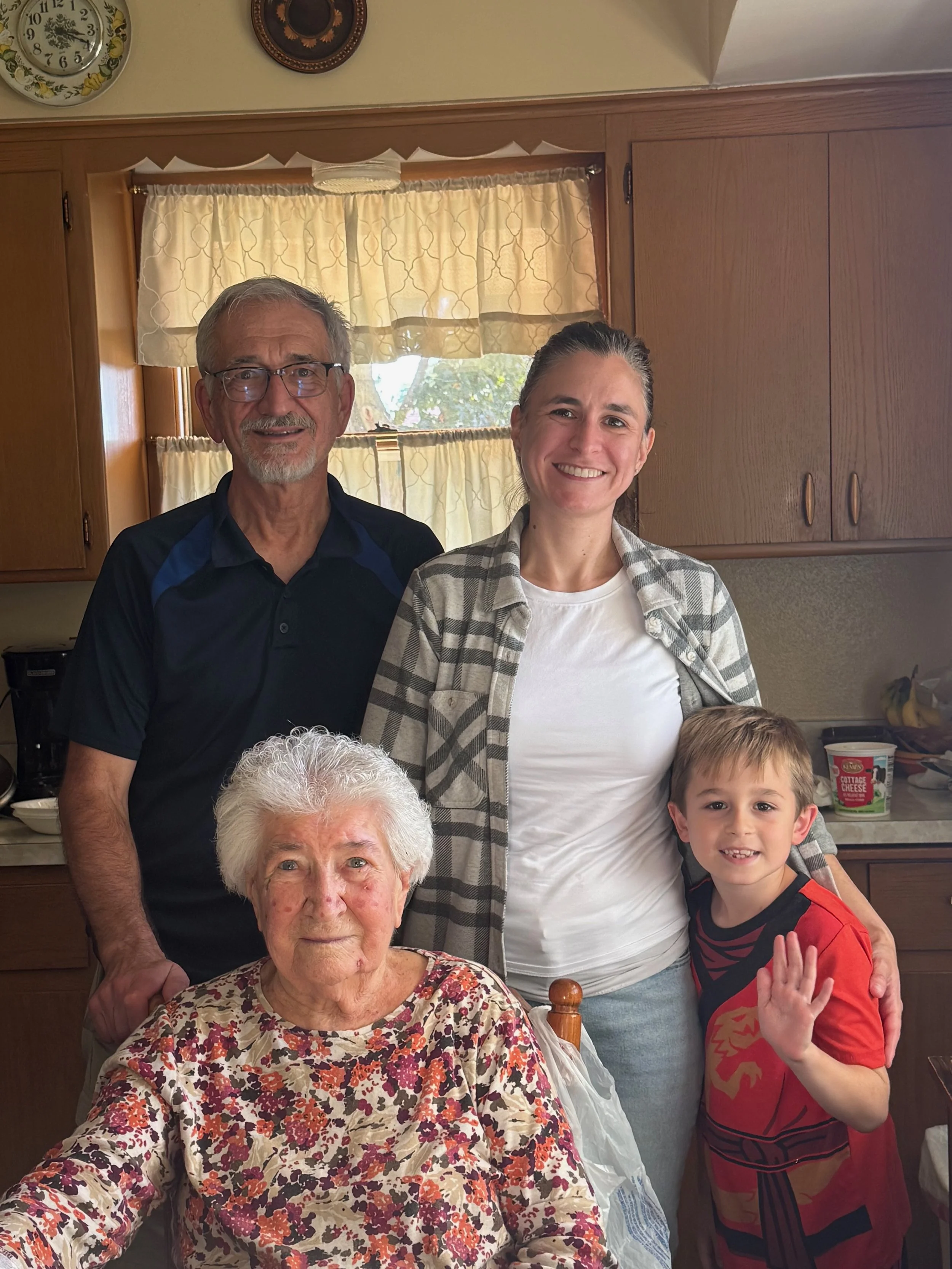 A multigenerational family gathering in a kitchen, with four people smiling and posing for a photo. An elderly woman in a floral blouse is seated in front, a middle-aged man with glasses and a beard is standing to her left, a middle-aged woman with long hair tied back is smiling and standing behind her, and a young boy in a red and black shirt is on the right, waving at the camera.