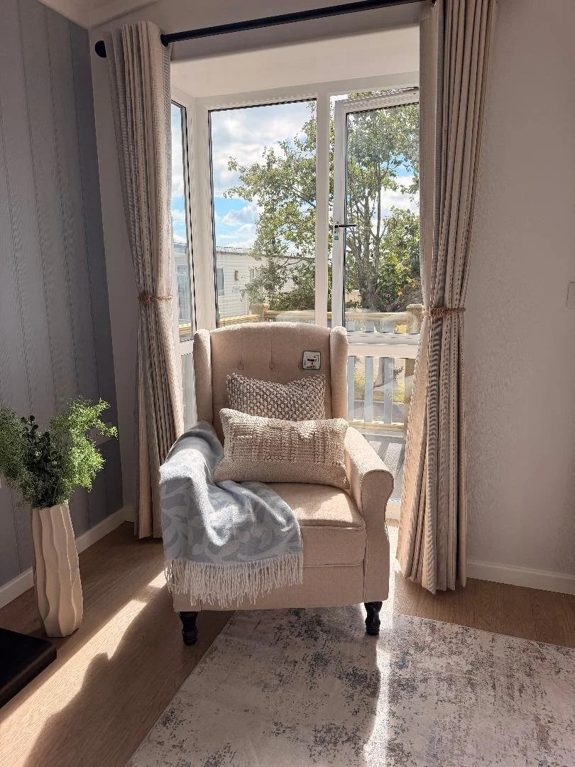 Sunlit corner of a room with a beige armchair, decorative pillows, a cozy throw, large window with sheer curtains, and view of trees and a building outside.