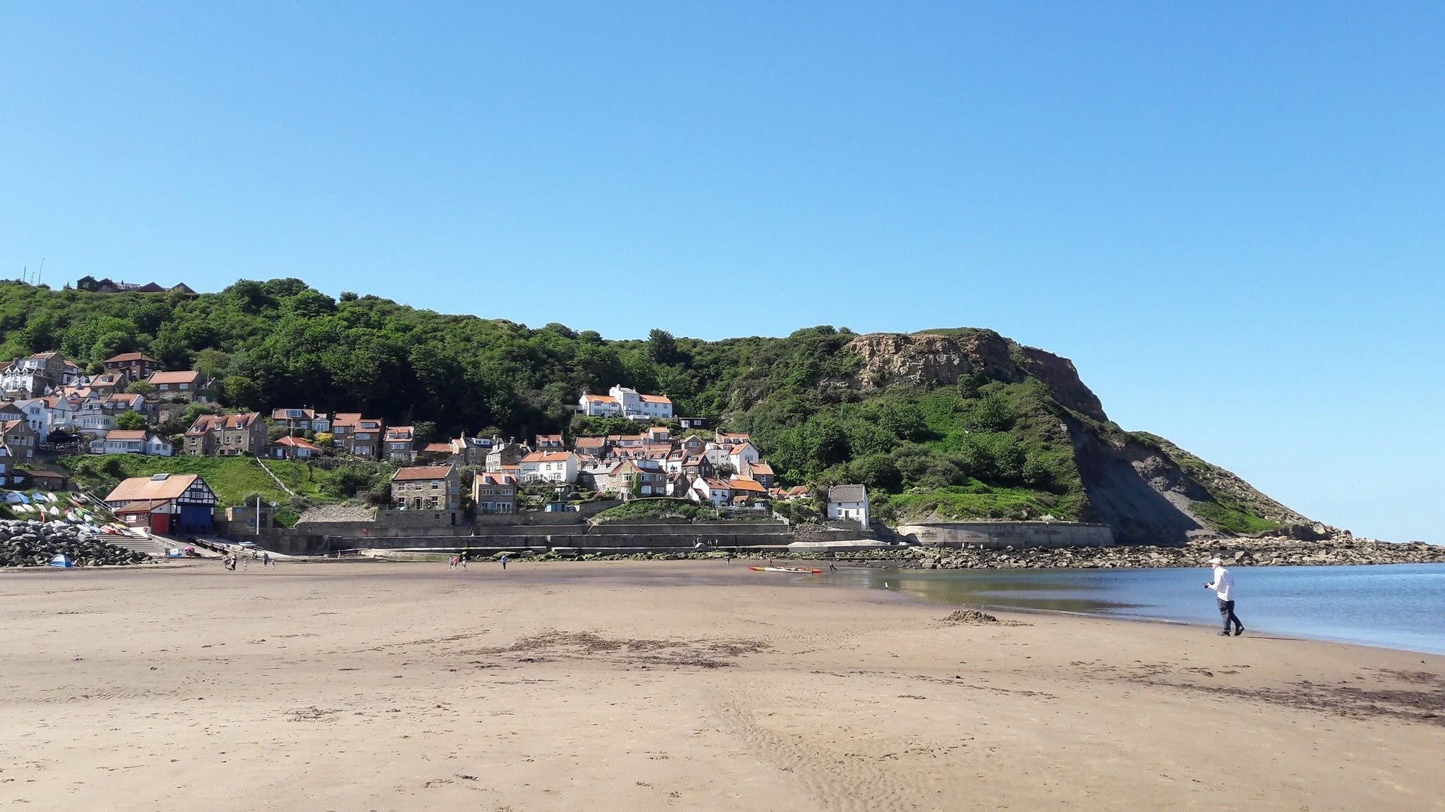 A beach with a sandy shore, a person walking near the water, and a hillside with houses and greenery under a clear blue sky.