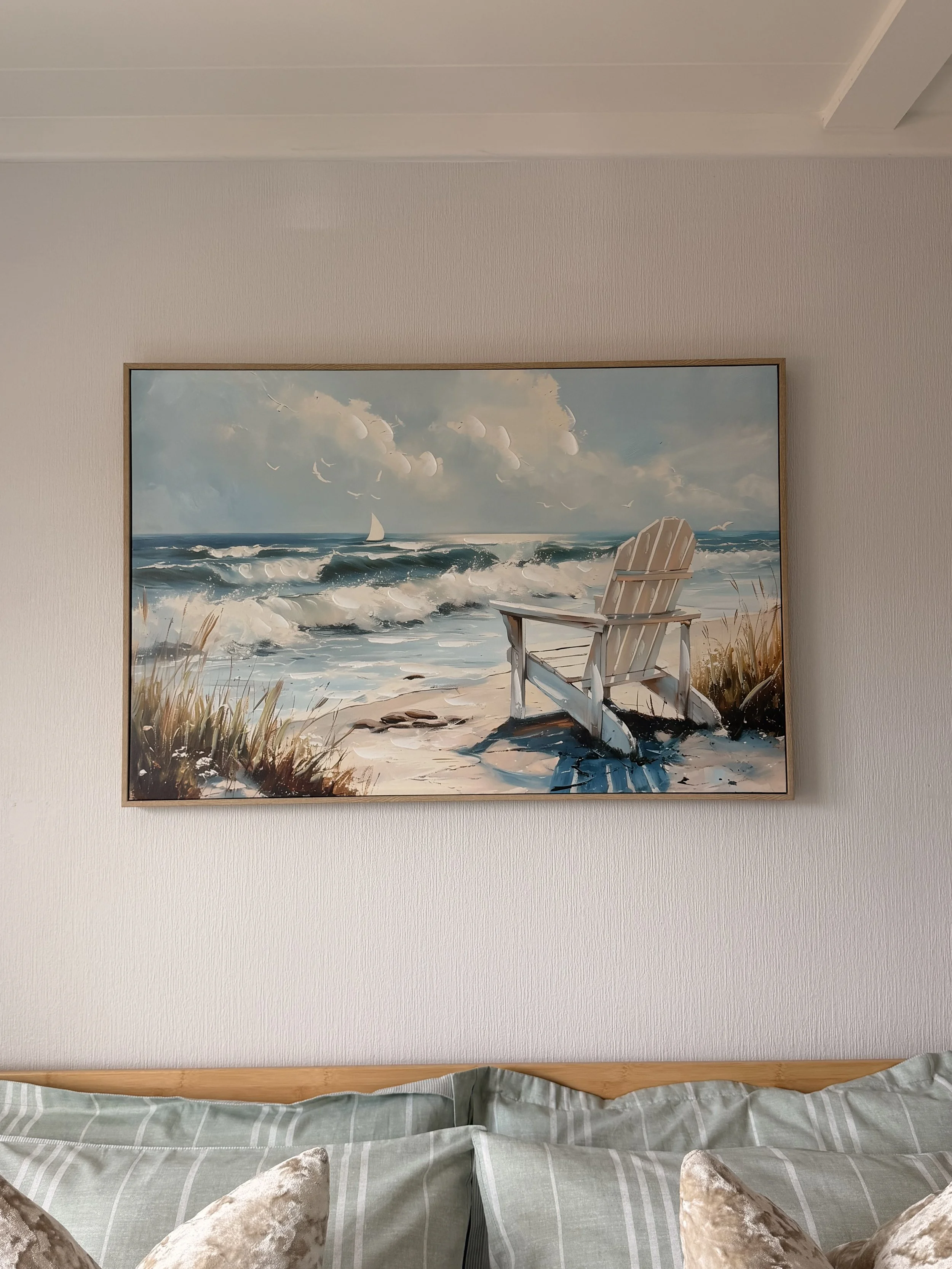Painting of a beach scene with waves, a sailboat, a cloudy sky, and a wooden beach chair, hung on a wall above a bed with striped pillows and a bedspread.