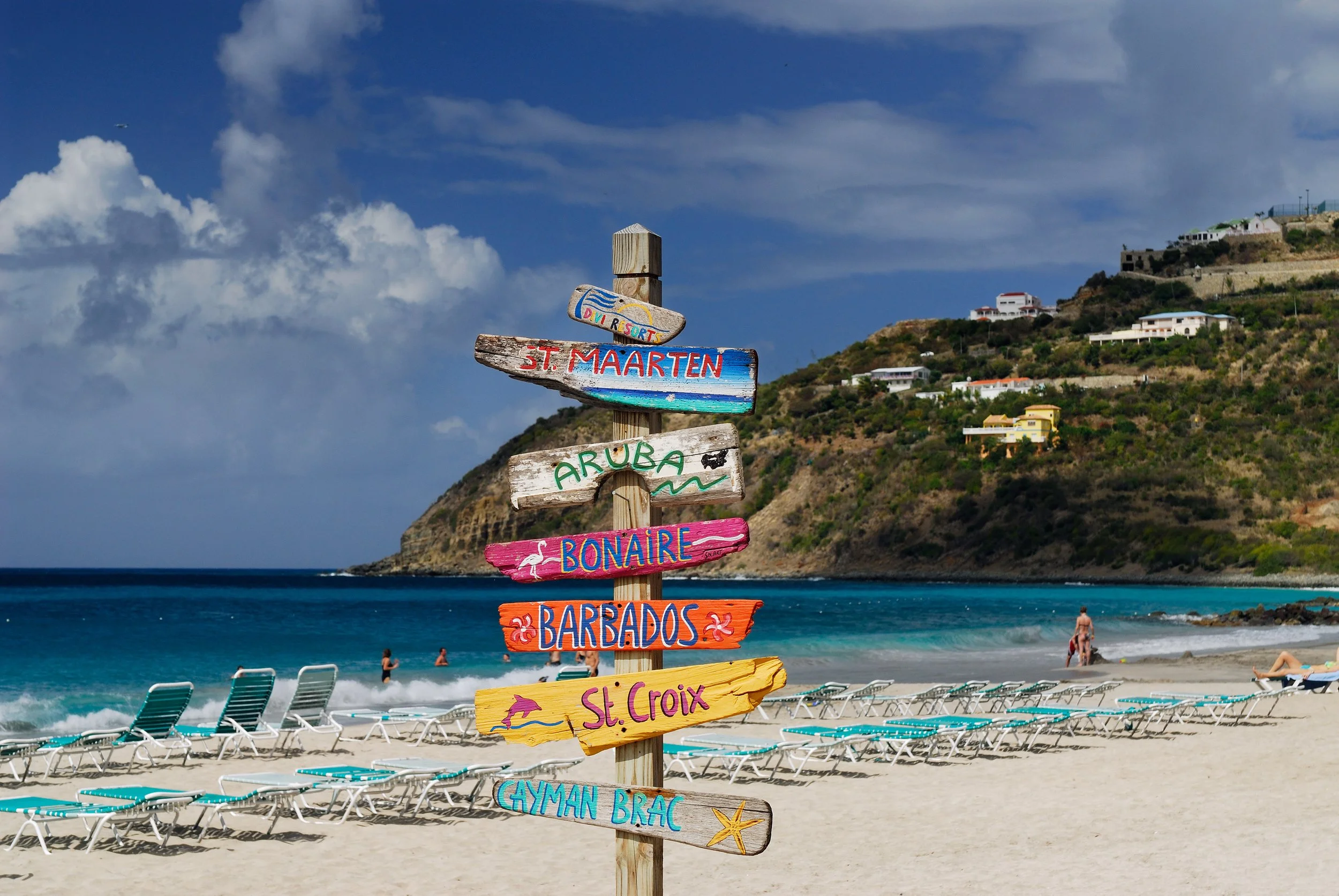 Colorful wooden signs on a beach with lounge chairs, ocean, and hillside houses in the background.