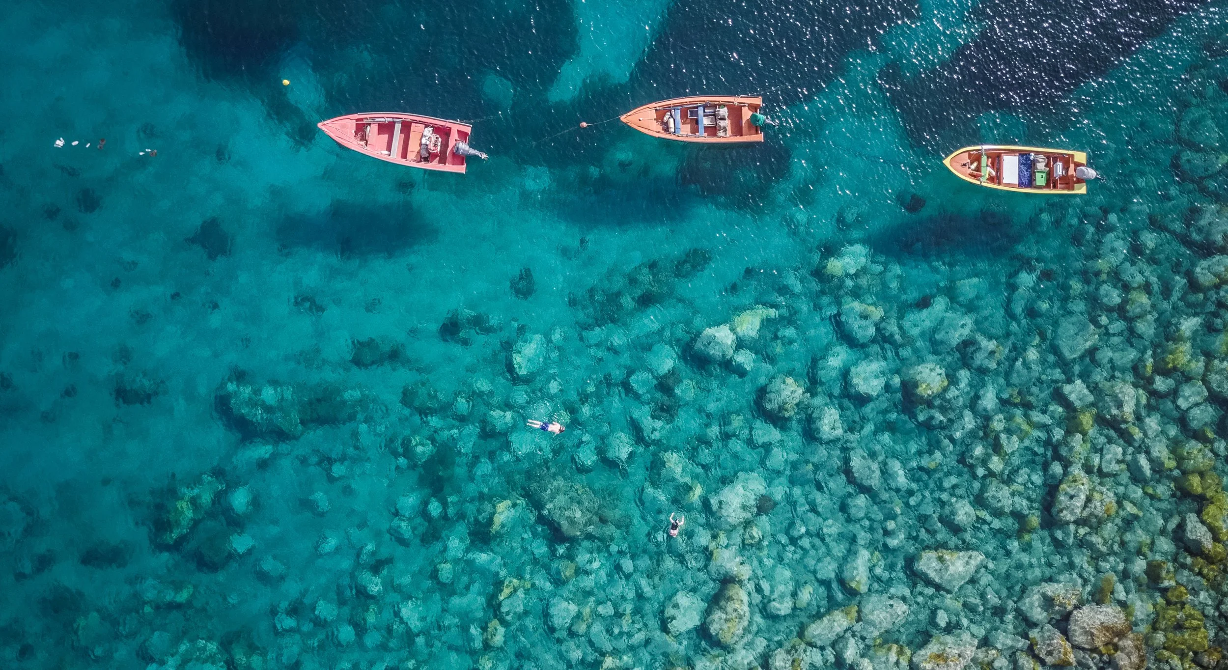 Four colorful boats anchored in clear, shallow ocean water with two people swimming nearby, and rocks visible beneath the water.
