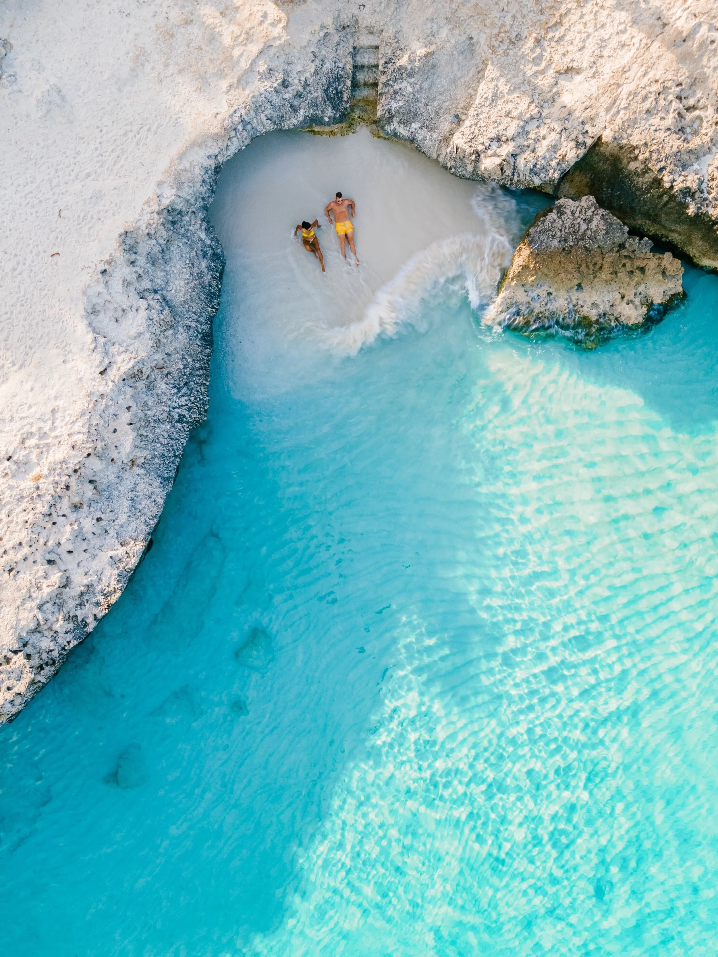 Two people lying on a sandy beach in a small cove with large rocks surrounding them, the water is clear and turquoise.