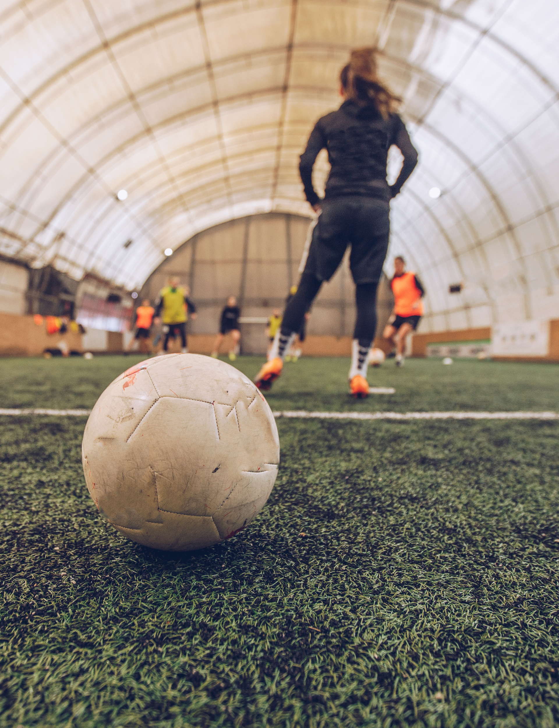 Soccer players practicing indoors on artificial turf with a soccer ball in the foreground