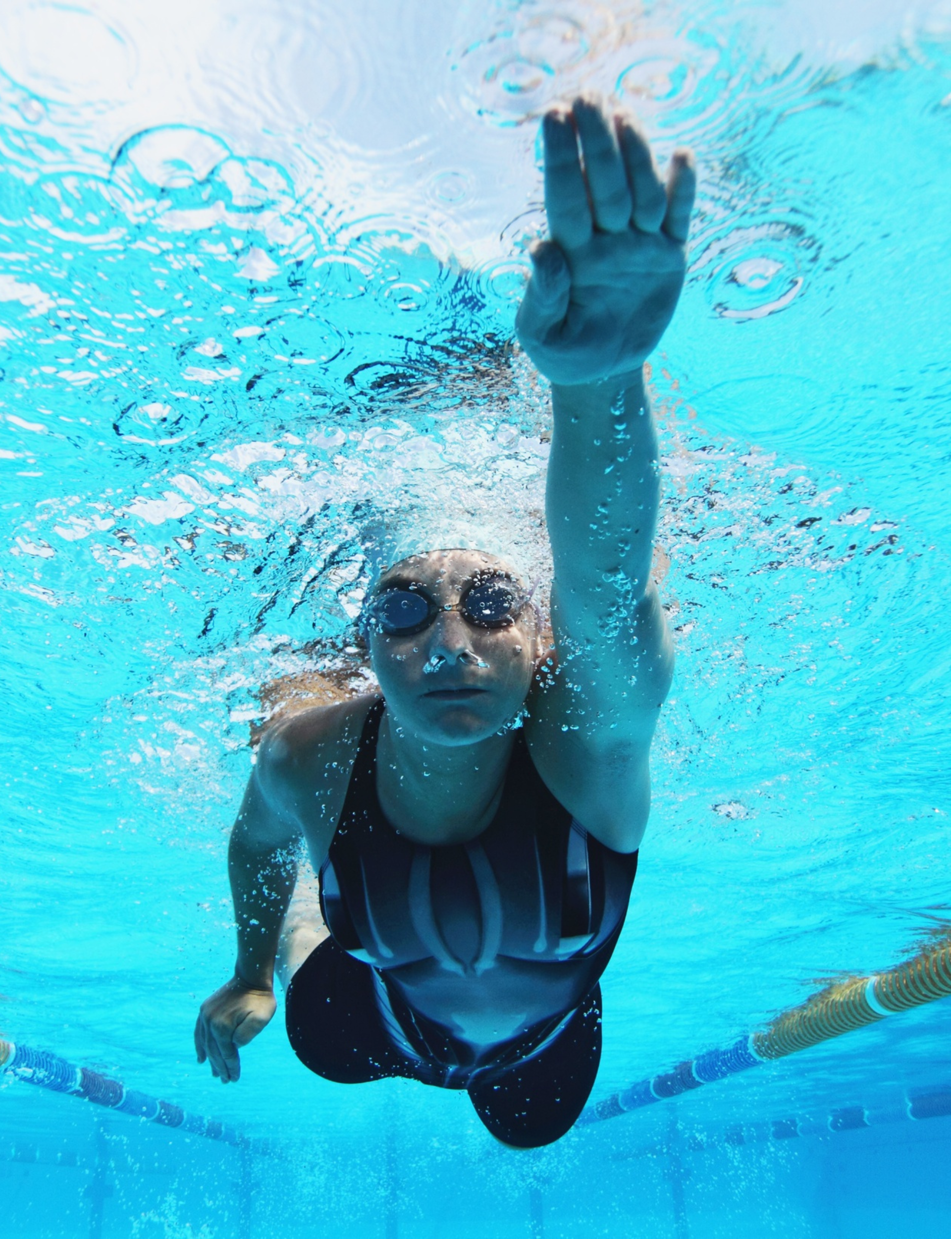 Swimmer wearing goggles and a black swimsuit reaching upward underwater in a swimming pool