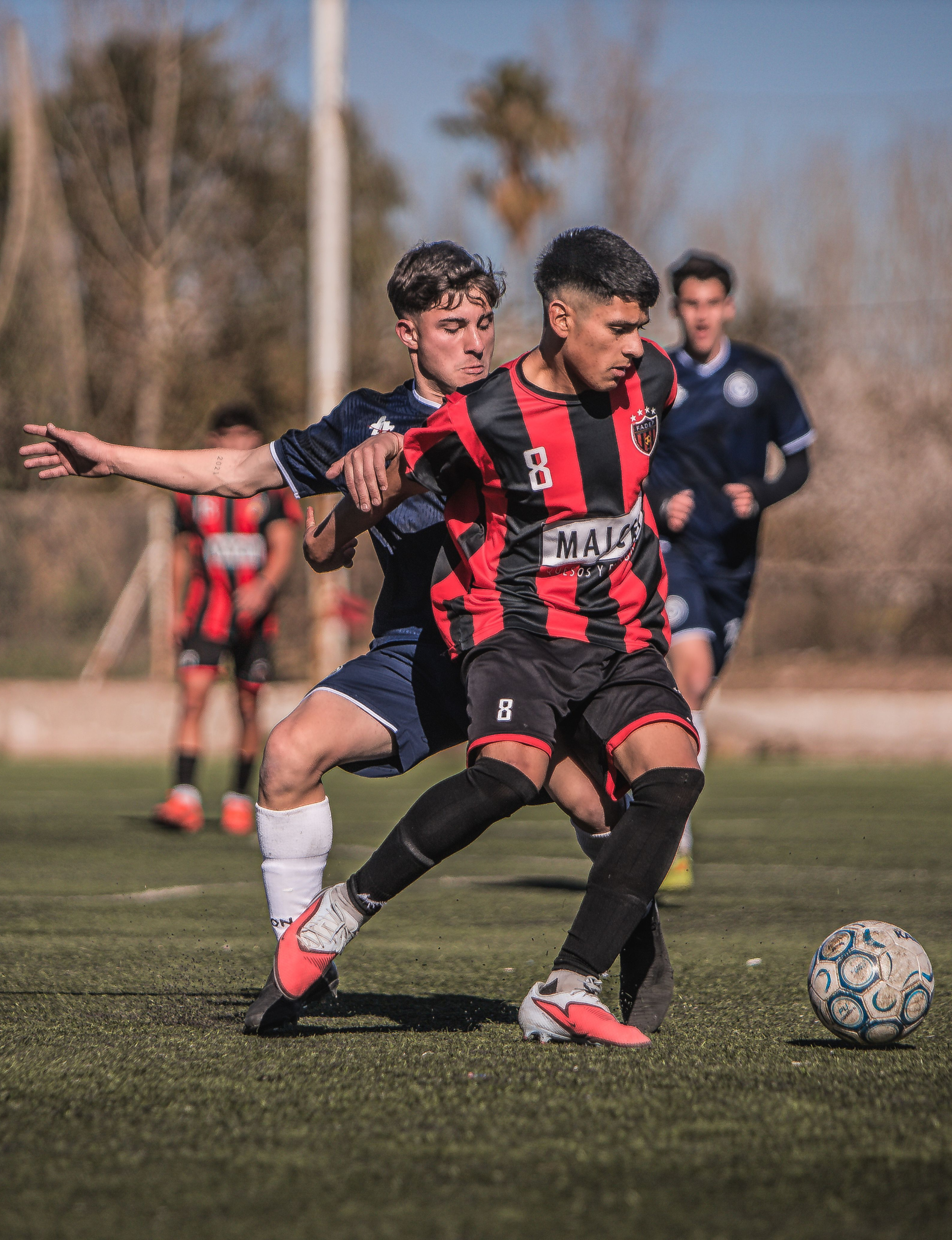 Soccer players competing for the ball on an outdoor field, with a player in a red-and-black No. 8 jersey in the foreground.