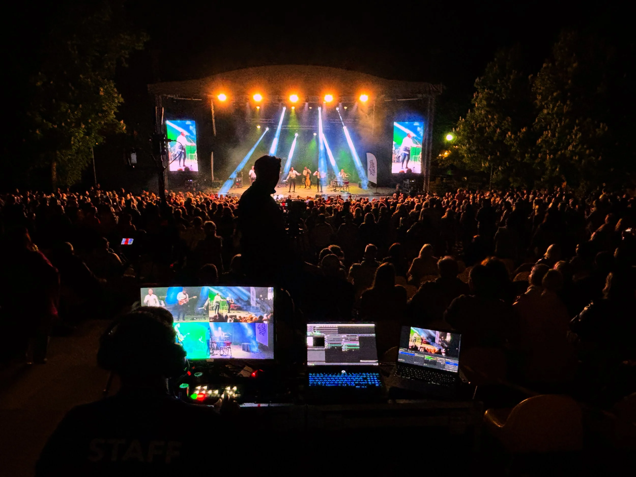 Nighttime outdoor concert with a large audience, stage with performers and bright spotlights, two large screens displaying the performance, and technical equipment in the foreground.