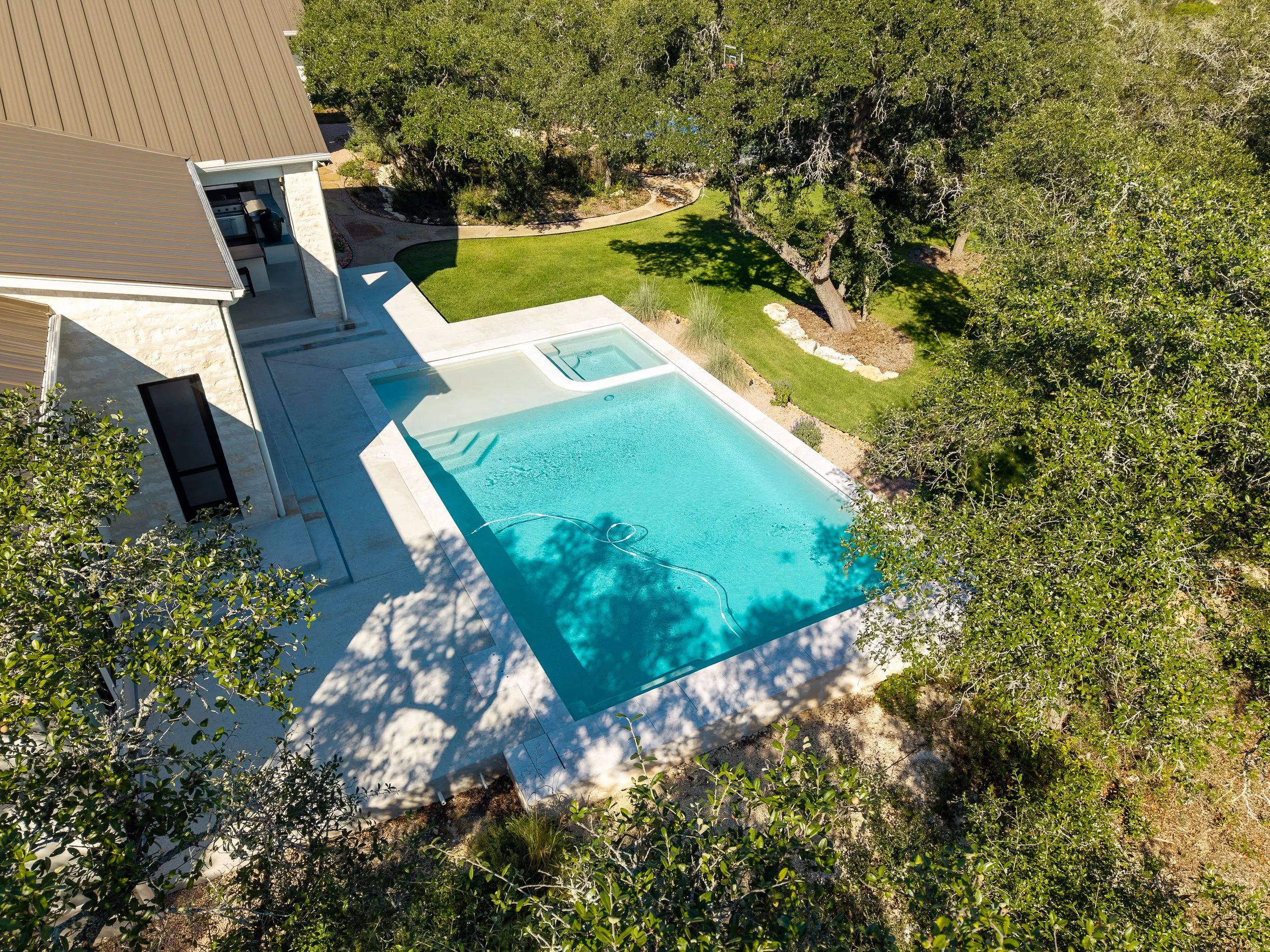 Aerial view of a landscaped backyard with a turquoise swimming pool, attached hot tub, stone patio, lawn, mature trees, and a house with an outdoor kitchen.