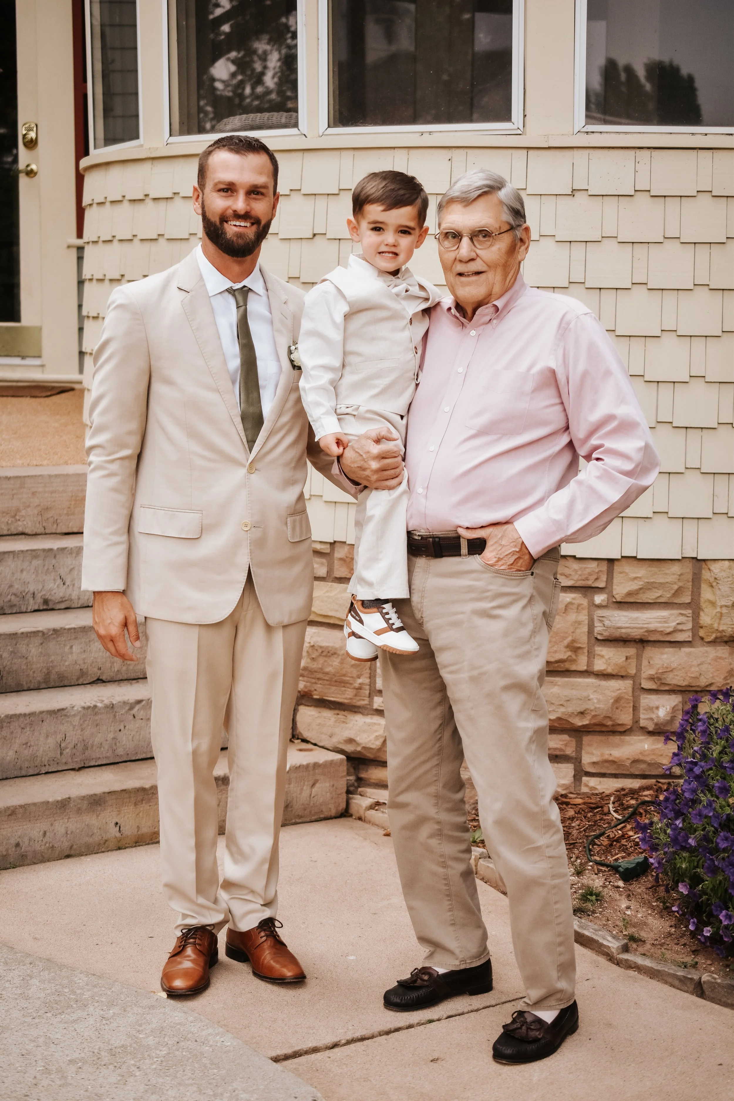 Smiling man in a suit, young boy, and older man posing together outside a house.