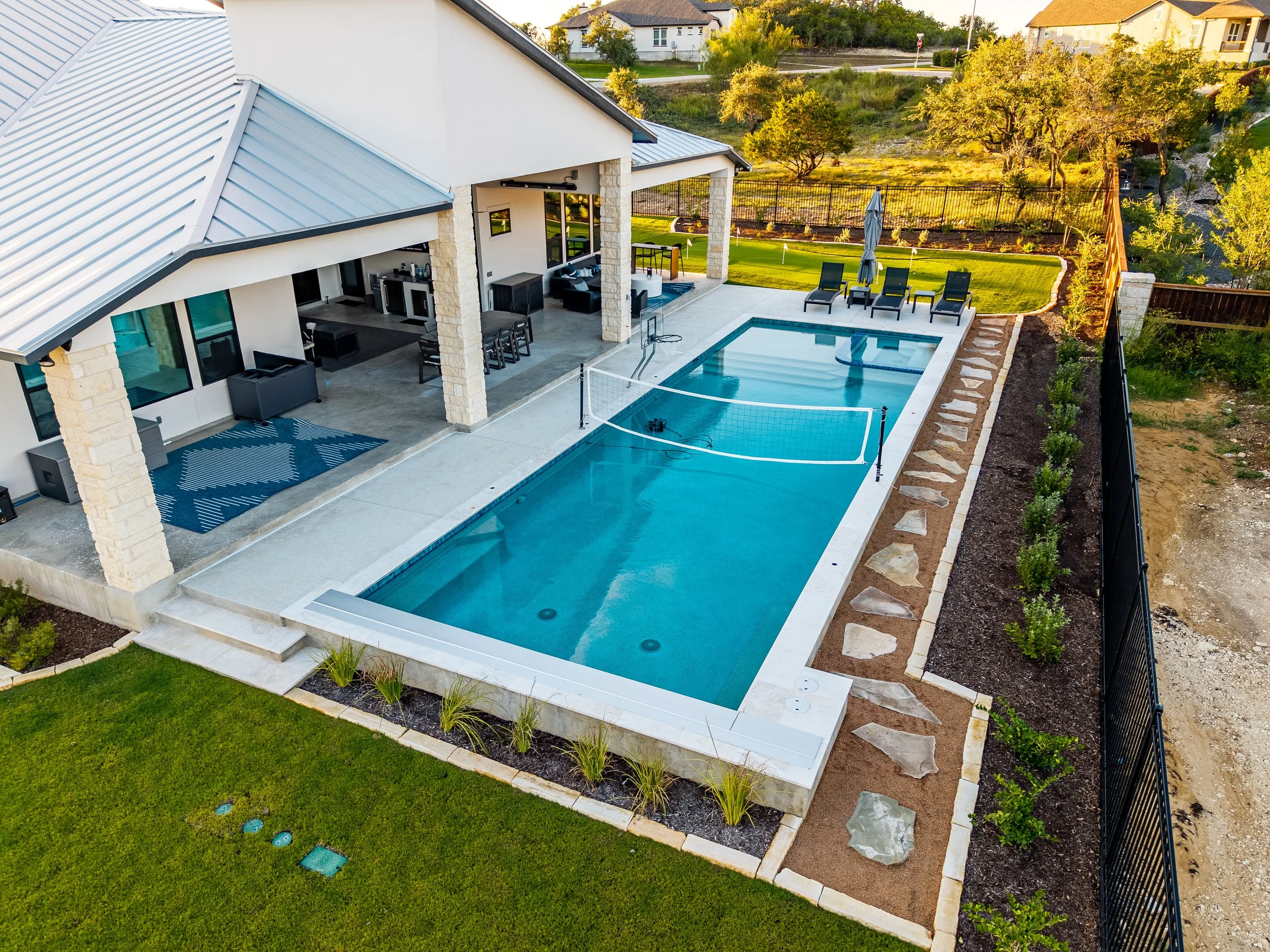 Modern backyard with a large swimming pool, volleyball net, covered outdoor kitchen and patio, lounge chairs, landscaped gardens, and manicured lawn