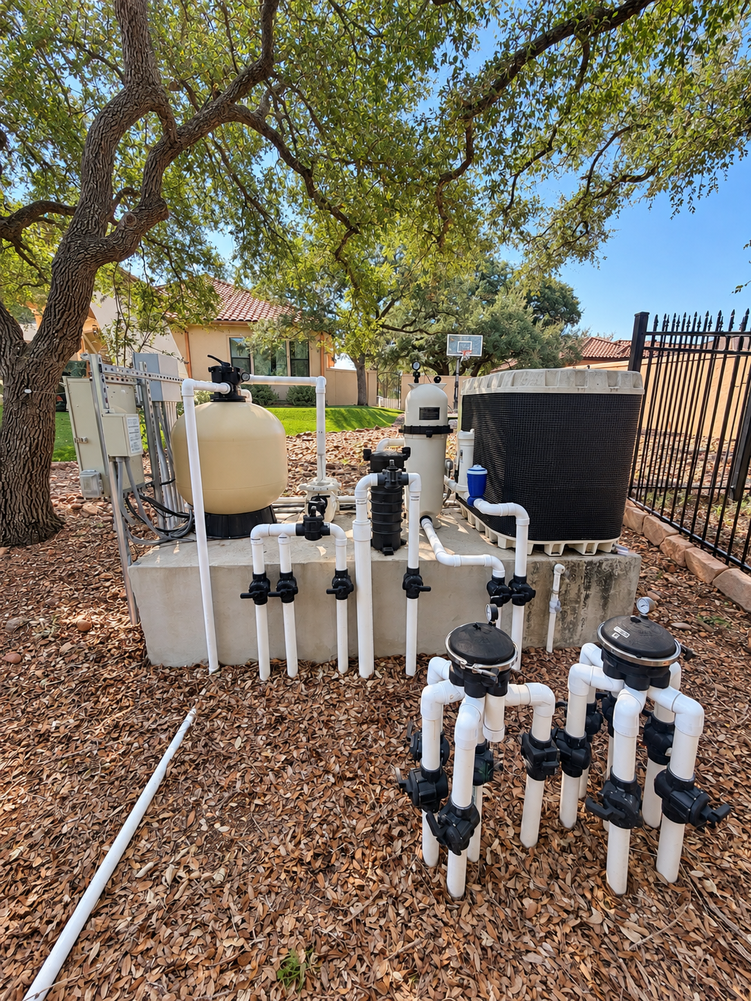 Residential swimming pool equipment with filters, pumps, white PVC pipes, and a large pool heater beside a fenced yard