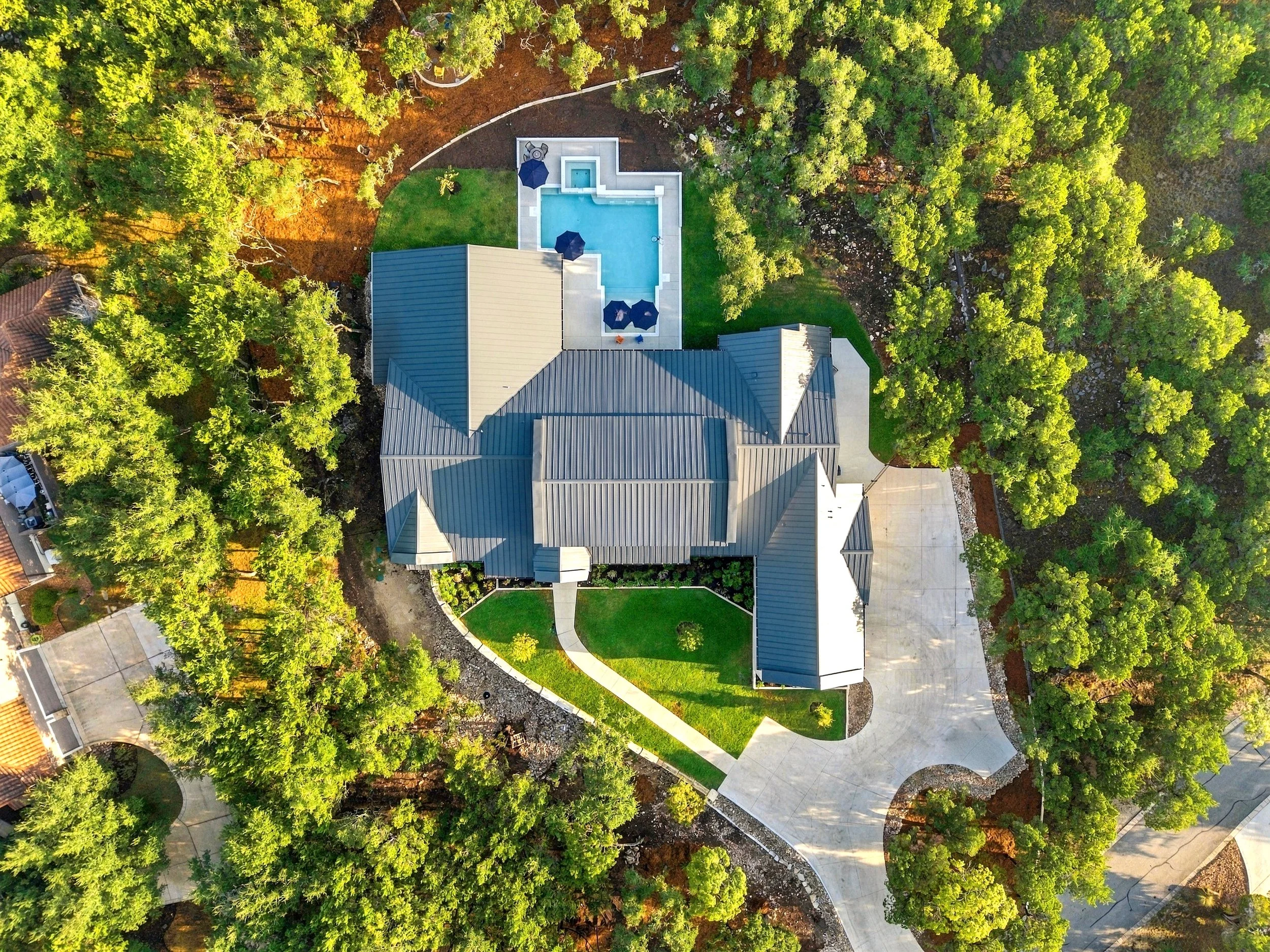 Aerial view of a large modern home with a swimming pool, landscaped yard, winding walkways, and surrounding trees.