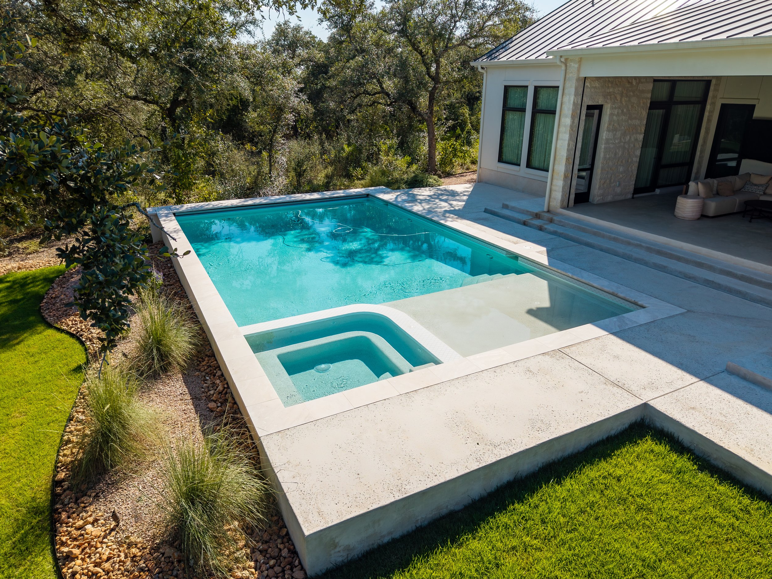 Modern home backyard with a turquoise swimming pool, attached spa, landscaped lawn, trees, and covered patio