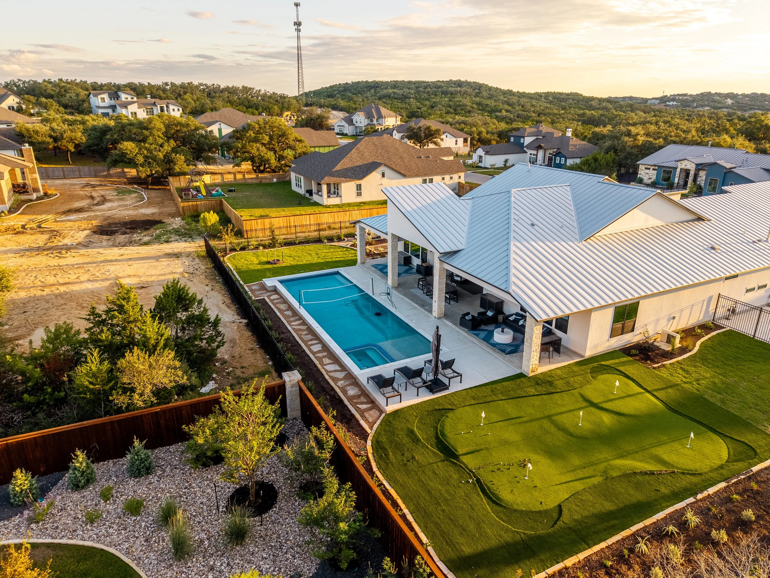 Aerial view of a luxury suburban home with a swimming pool, covered patio, backyard putting green, and wooded hills at sunset.