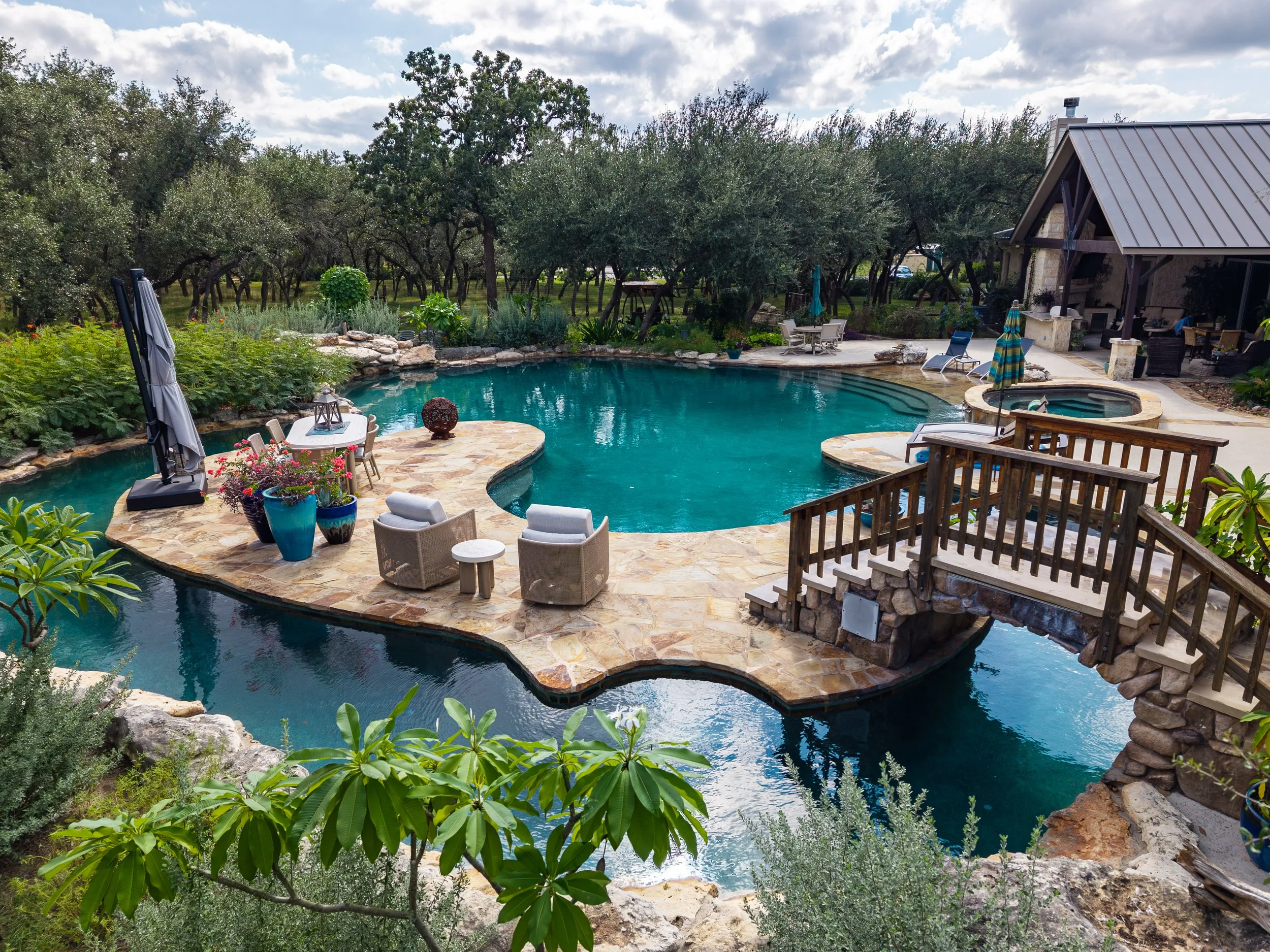 Luxurious backyard pool with stone patio, hot tub, lounge chairs, umbrellas, lush landscaping, and a poolside cabana