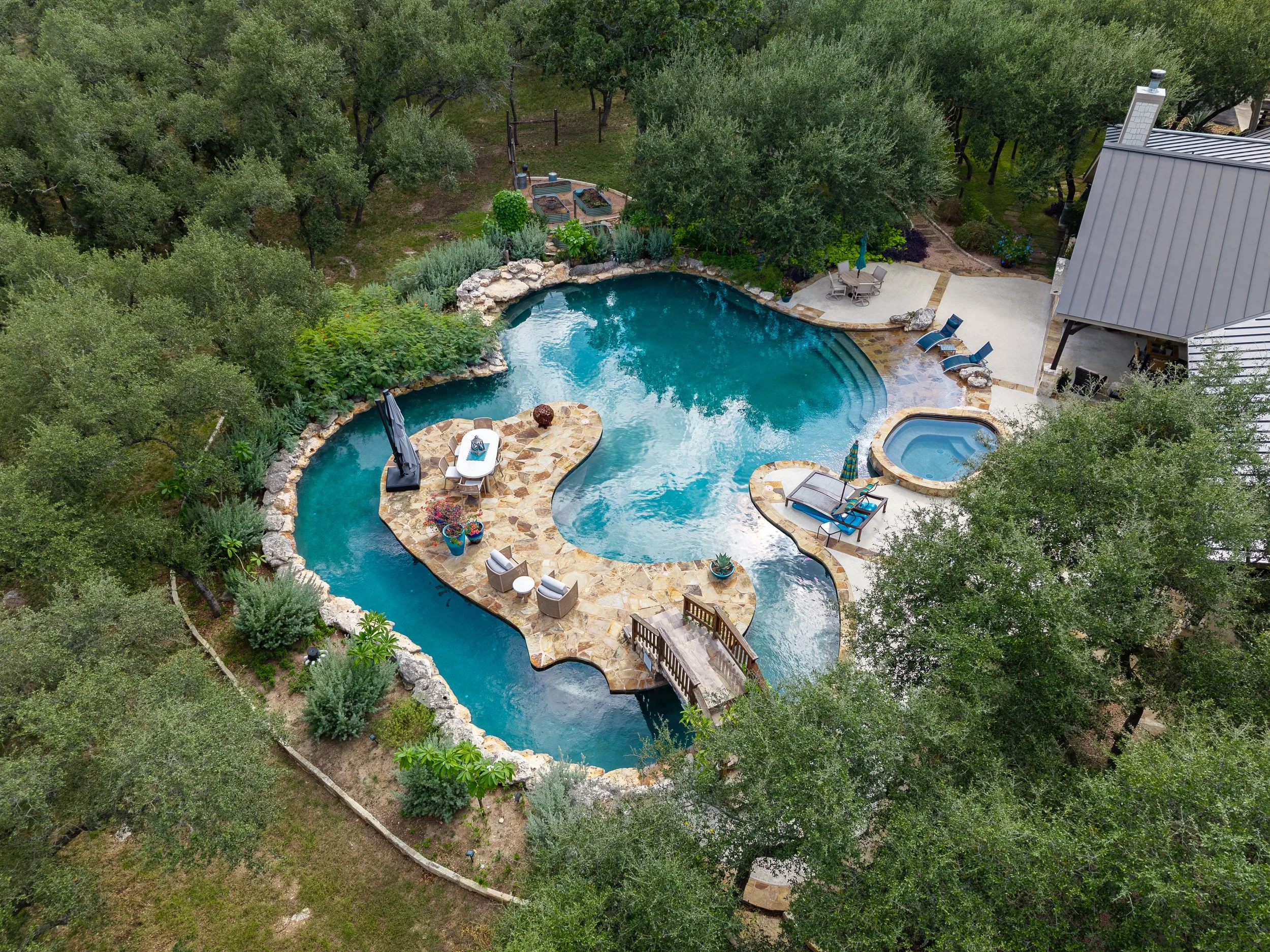 Aerial view of a resort-style swimming pool with a spa, stone patios, lounge chairs, gardens, and surrounding olive trees.