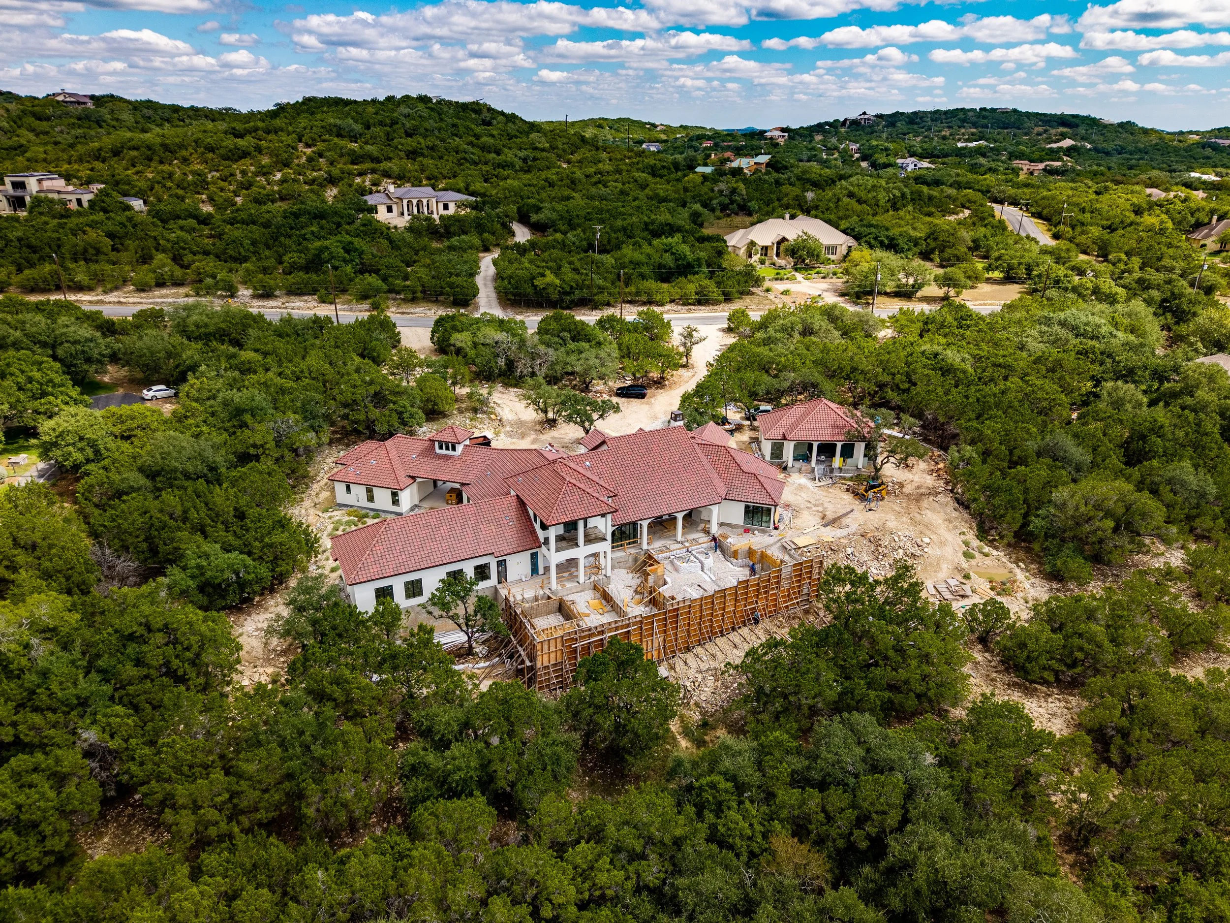 Aerial view of a large red-tile-roof home under construction in a wooded residential neighborhood with rolling hills and scattered houses.