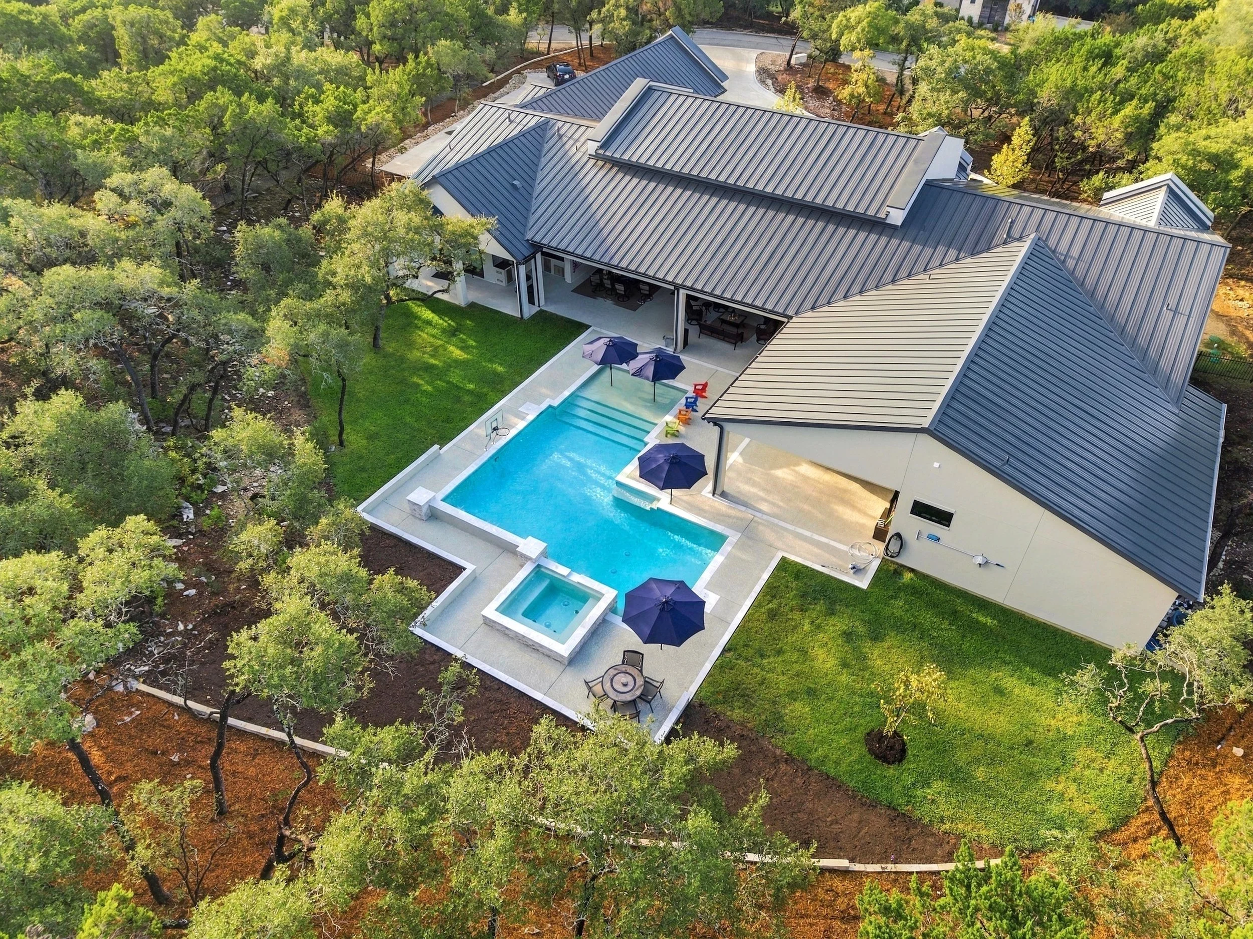 Aerial view of a modern home with a gray metal roof, swimming pool, hot tub, patio, and landscaped wooded yard