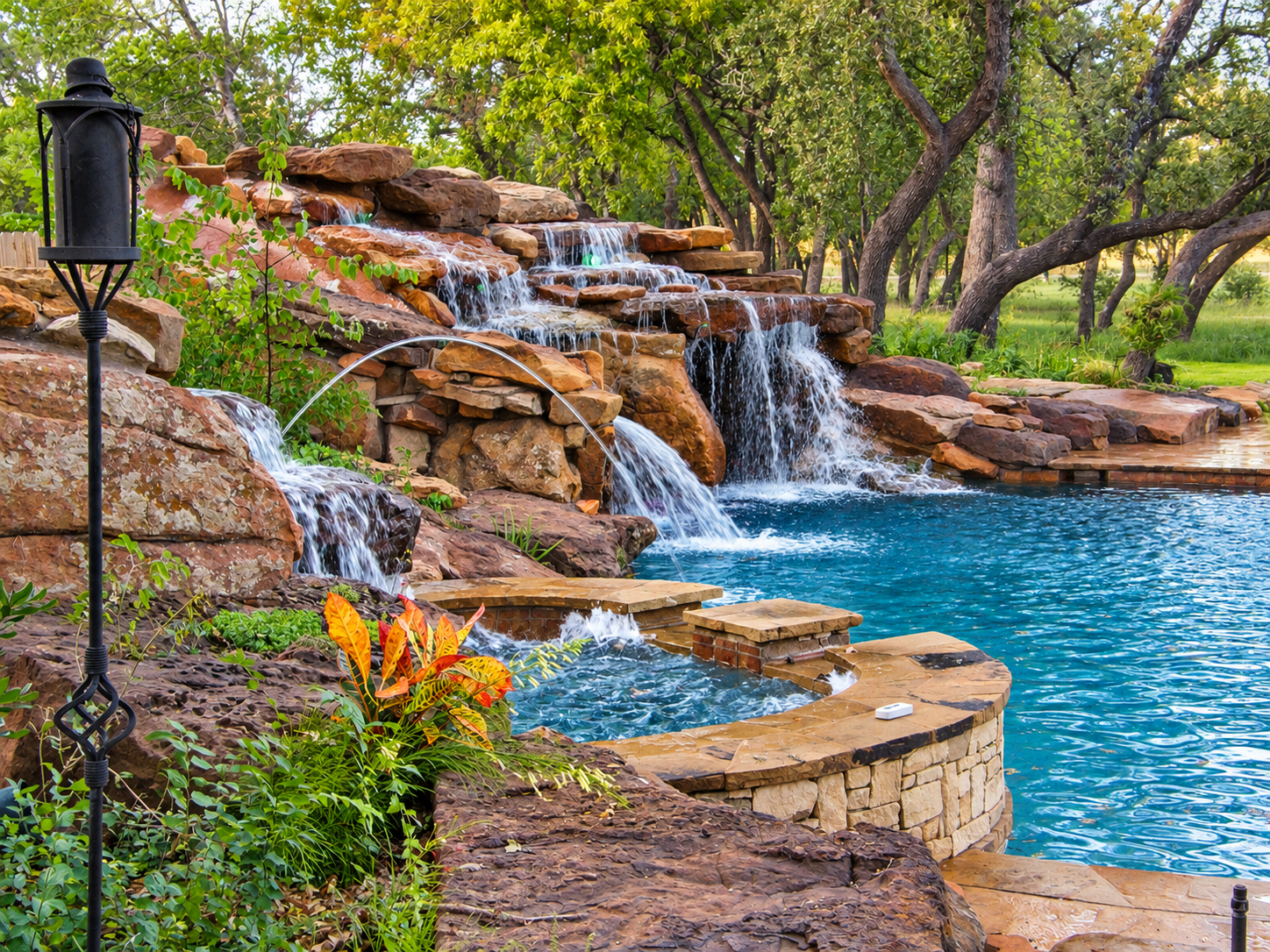 Luxury backyard pool with multiple rock waterfalls, spa, lush landscaping, and outdoor lantern