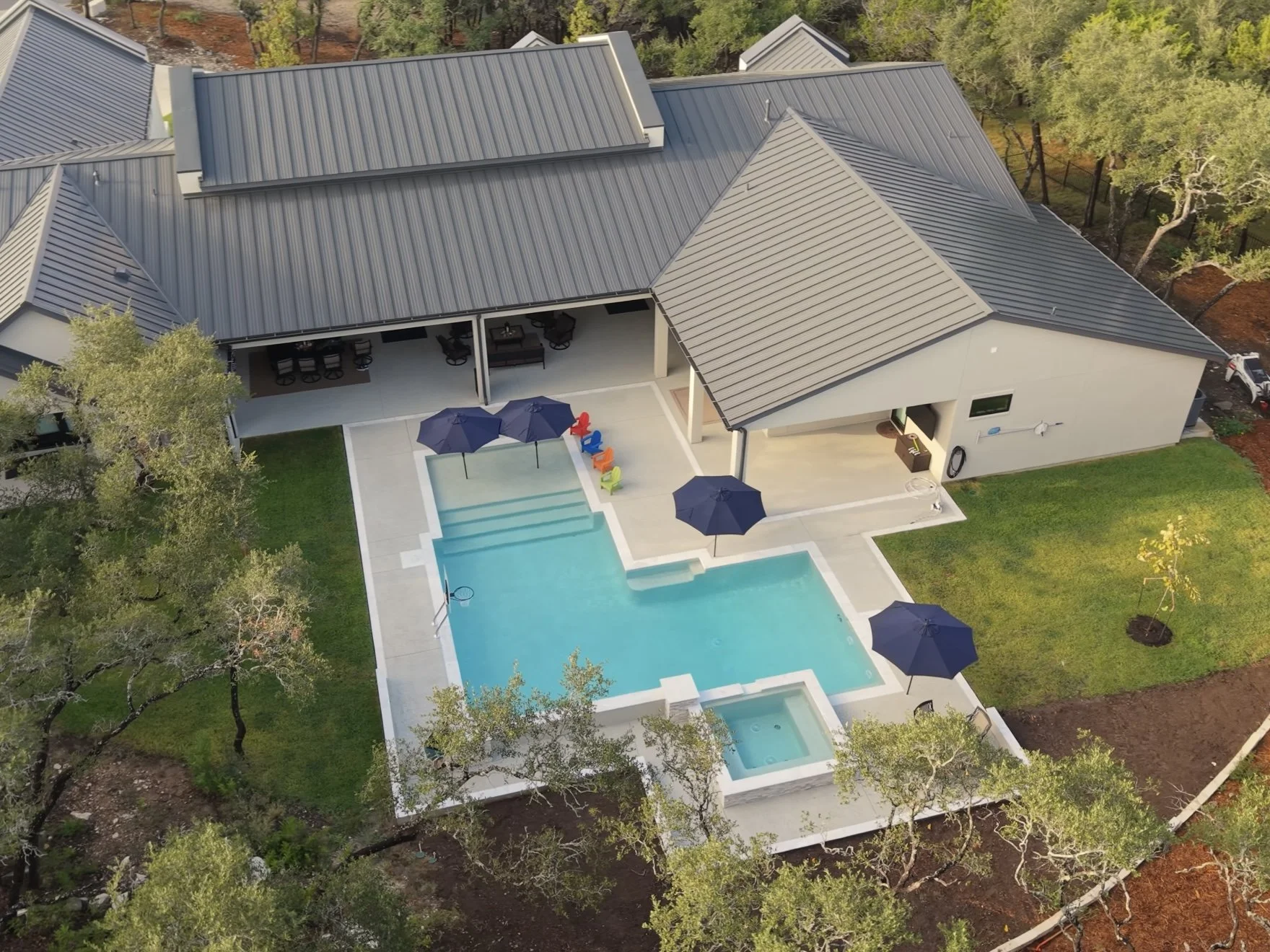 Aerial view of a modern home with gray metal roofs, covered patio, swimming pool, hot tub, and outdoor umbrellas.