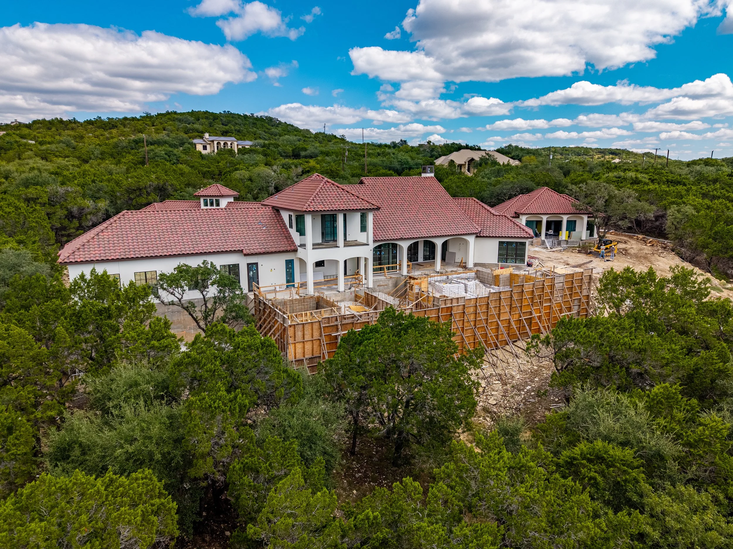 Aerial view of a large Mediterranean-style home under construction with red tile roofs, surrounded by dense greenery under a bright blue sky