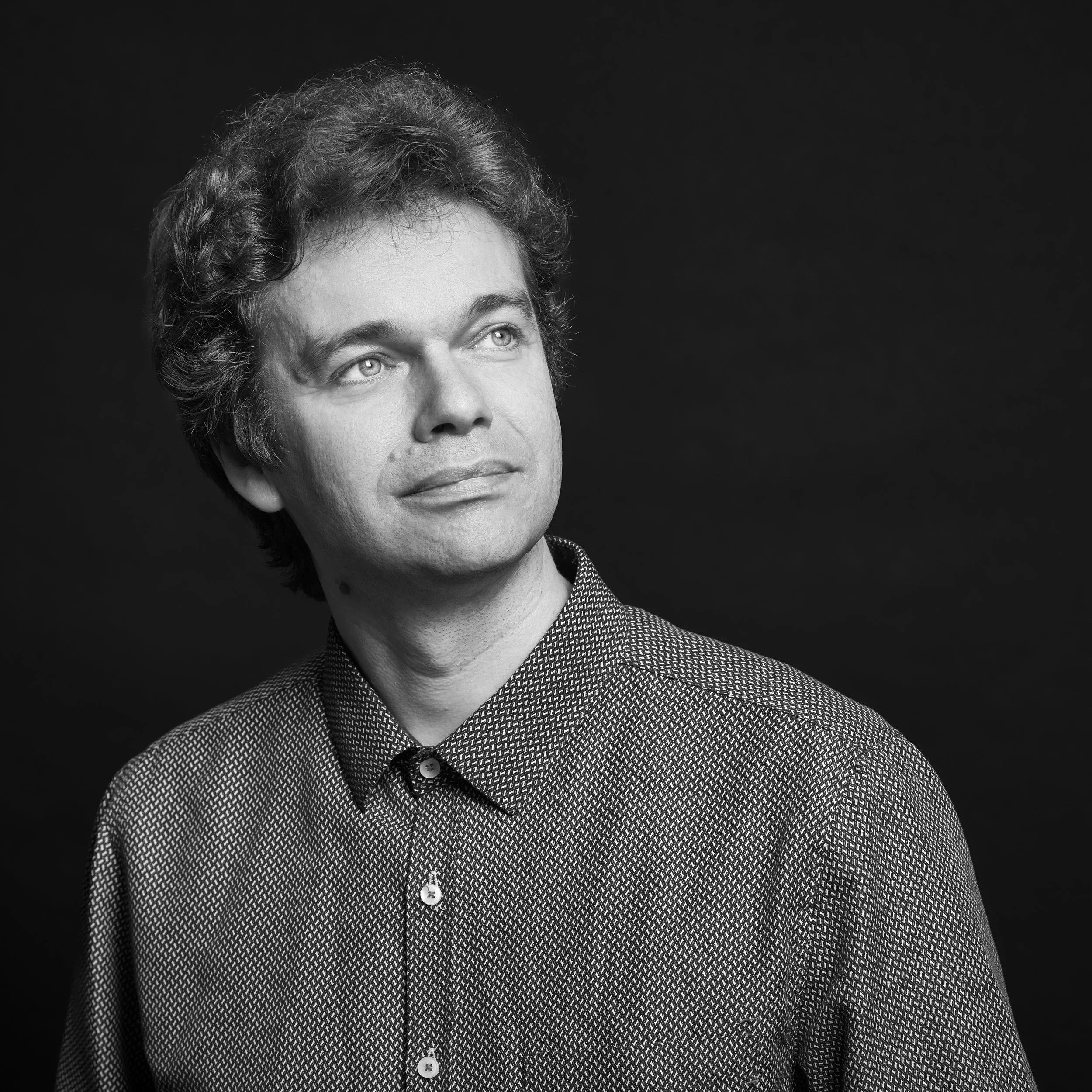 Un portrait en noir et blanc d'un homme avec des cheveux bouclés et un sourire léger, portant une veste à motifs avec une chemise en dessous, contre un fond noir.