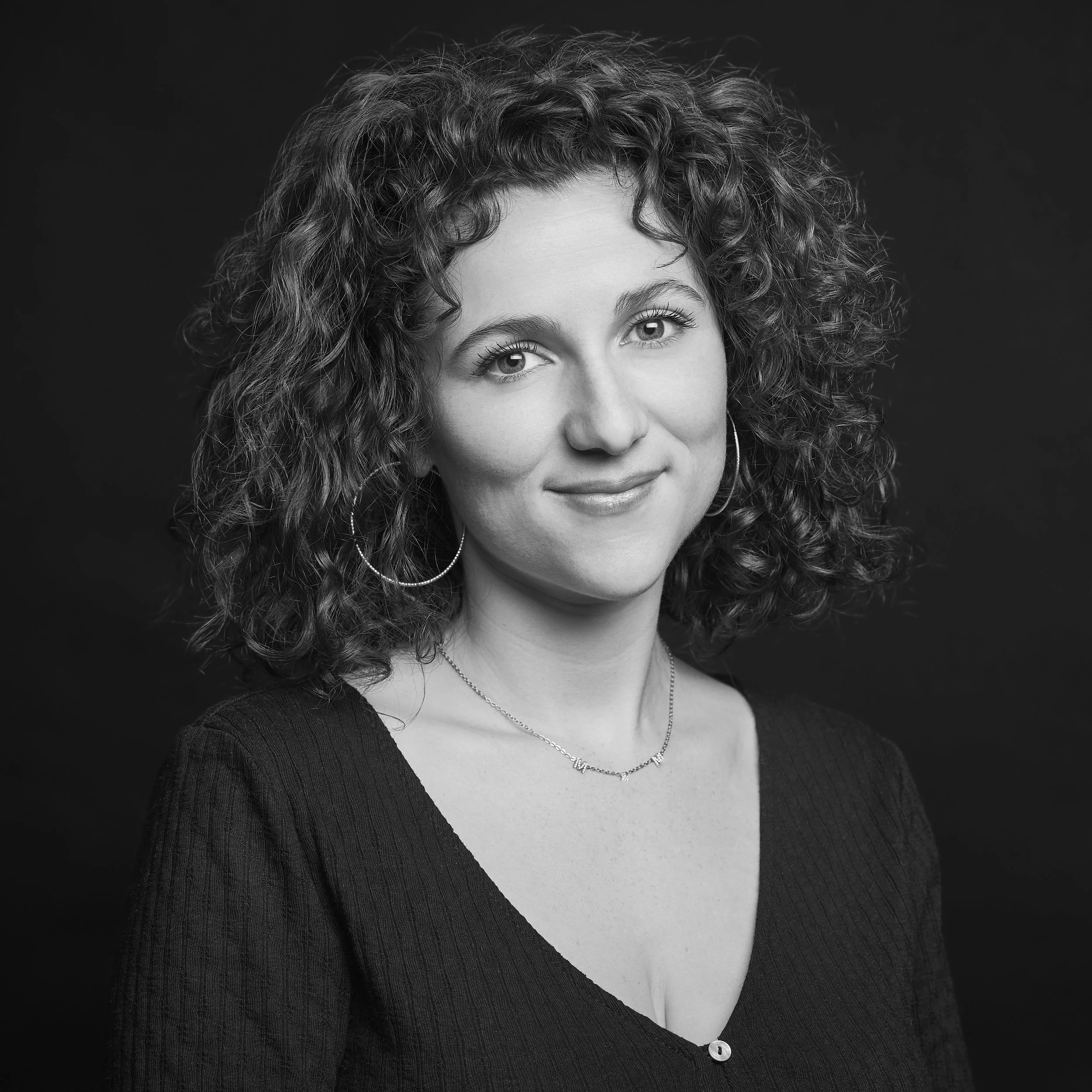 Portrait d'une femme avec des cheveux bouclés, portant des boucles d'oreilles et un collier, photographiée en noir et blanc contre un fond sombre.