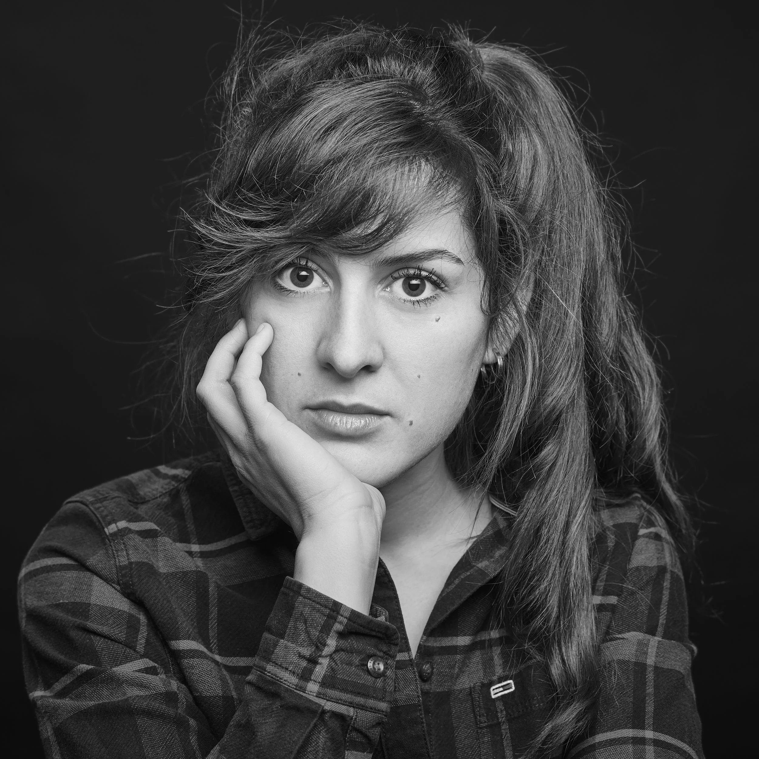 Portrait d'une femme avec des cheveux bouclés et longs, portant une veste à carreaux. La photo est en noir et blanc et sa main repose sur son visage, avec son index proche de son œil.