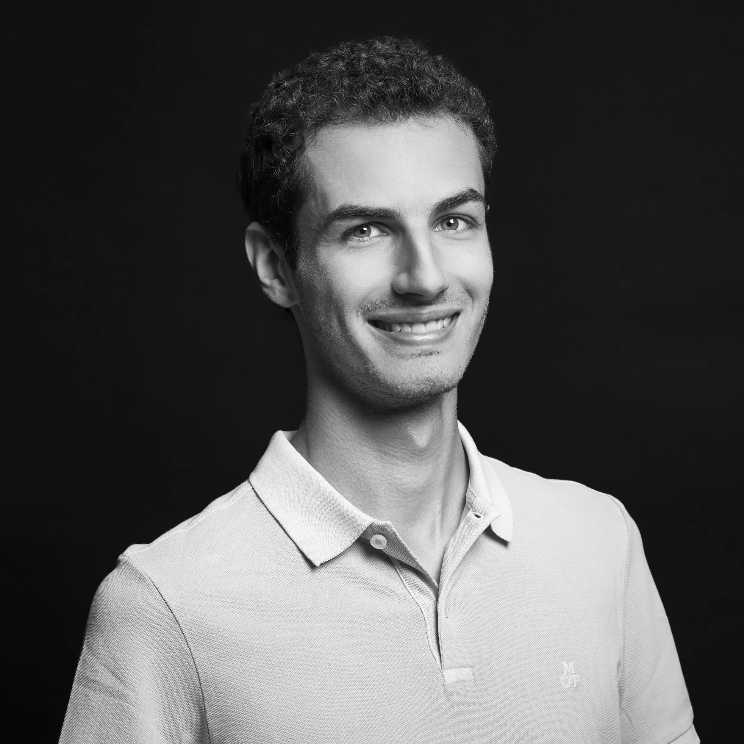 Portrait en noir et blanc d'un homme souriant, portant une chemise à col boutonné, avec un fond noir uniforme.