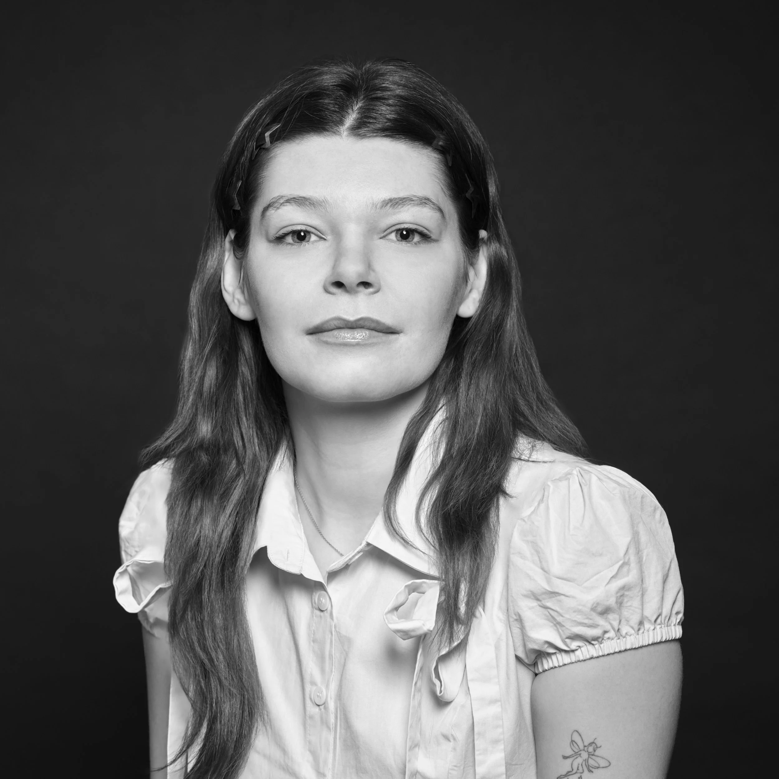 Portrait d'une femme avec cheveux longs, porteur d'une chemise blanche, expression neutre, fond noir.