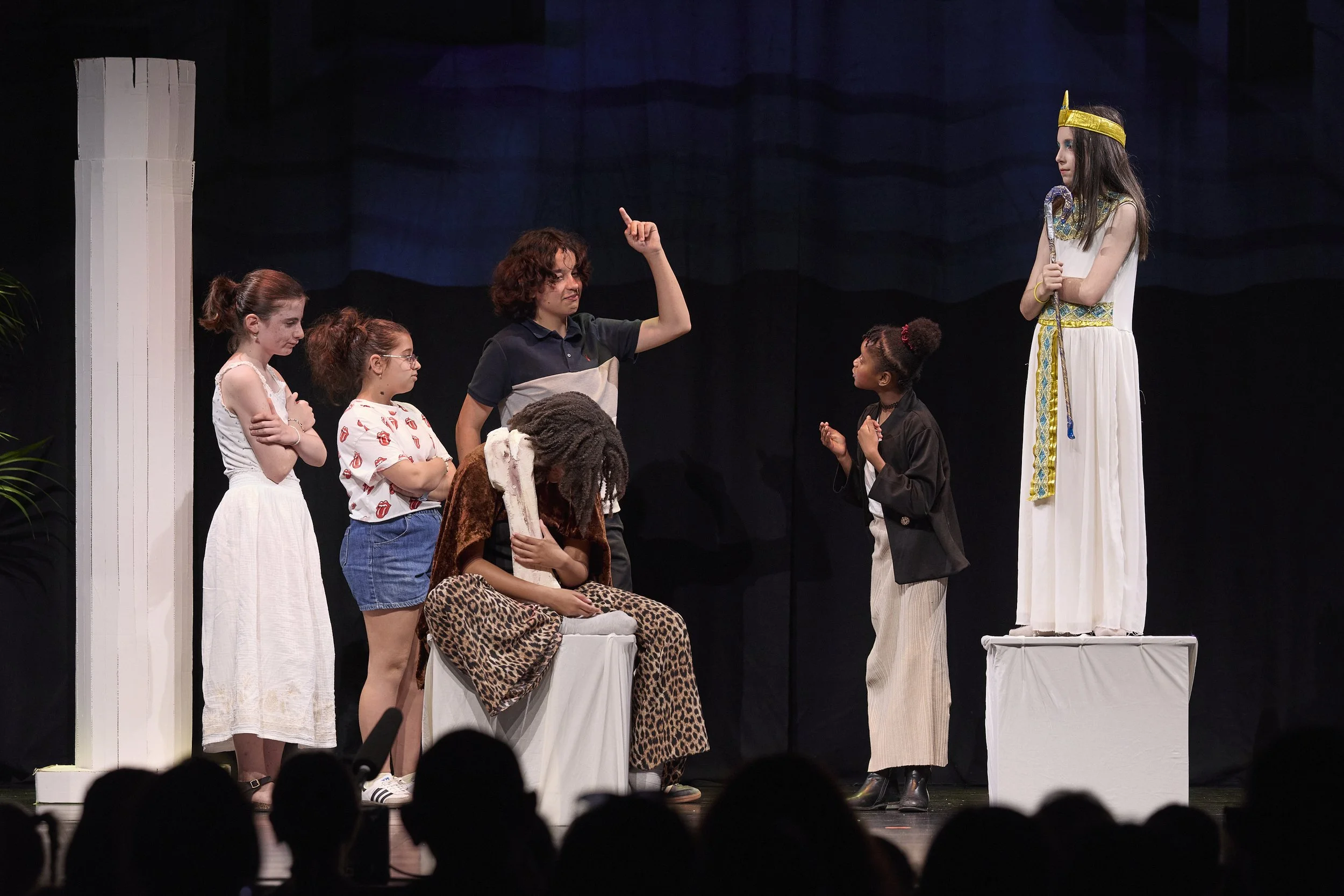 Scène de théâtre réunissant plusieurs jeunes acteurs, dont une reine vêtue de blanc et d’or, devant un décor sombre.