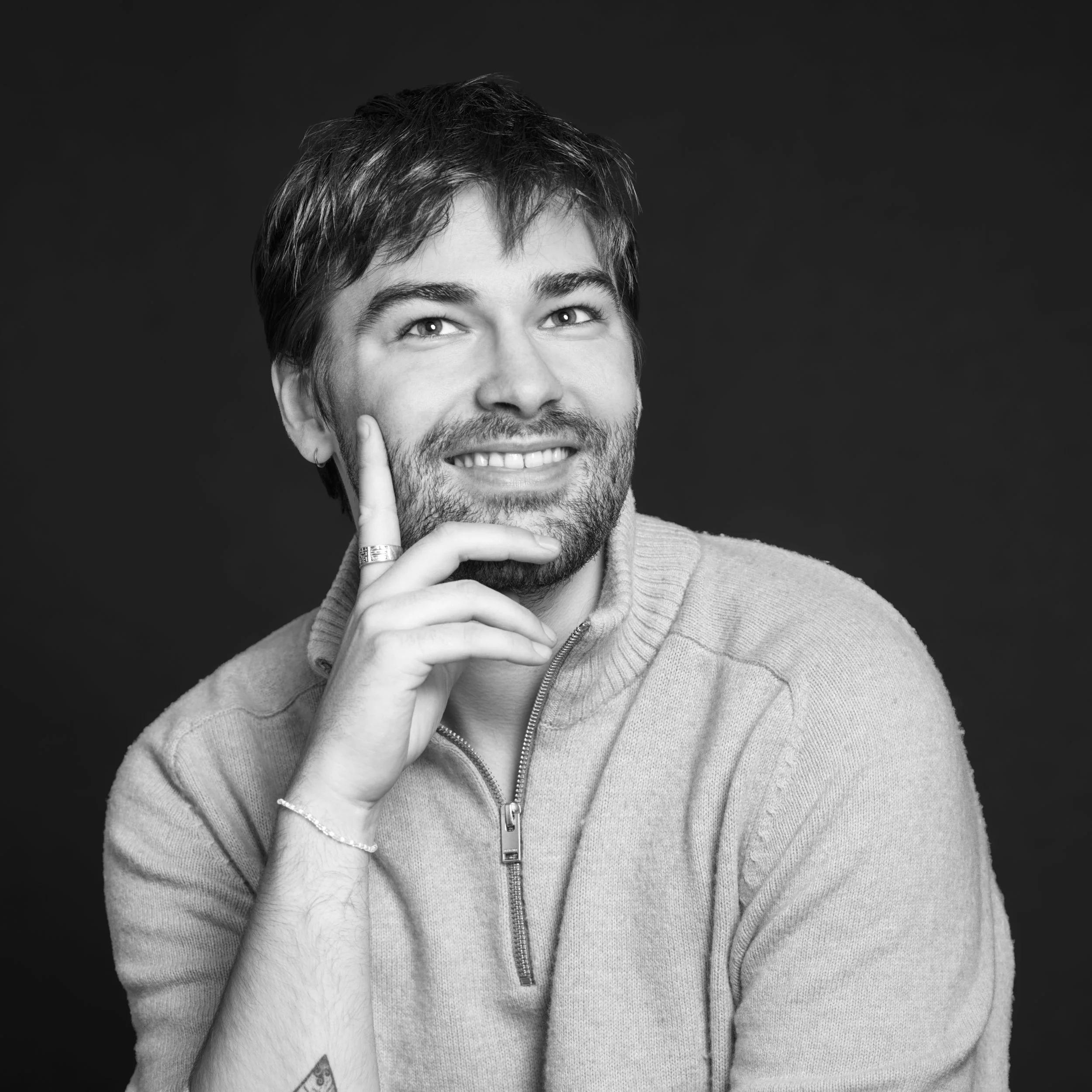 Un homme souriant avec une barbe, touchant son visage avec son doigt, porteur de bijoux, portant un pull gris avec une fermeture éclair, sur fond noir.