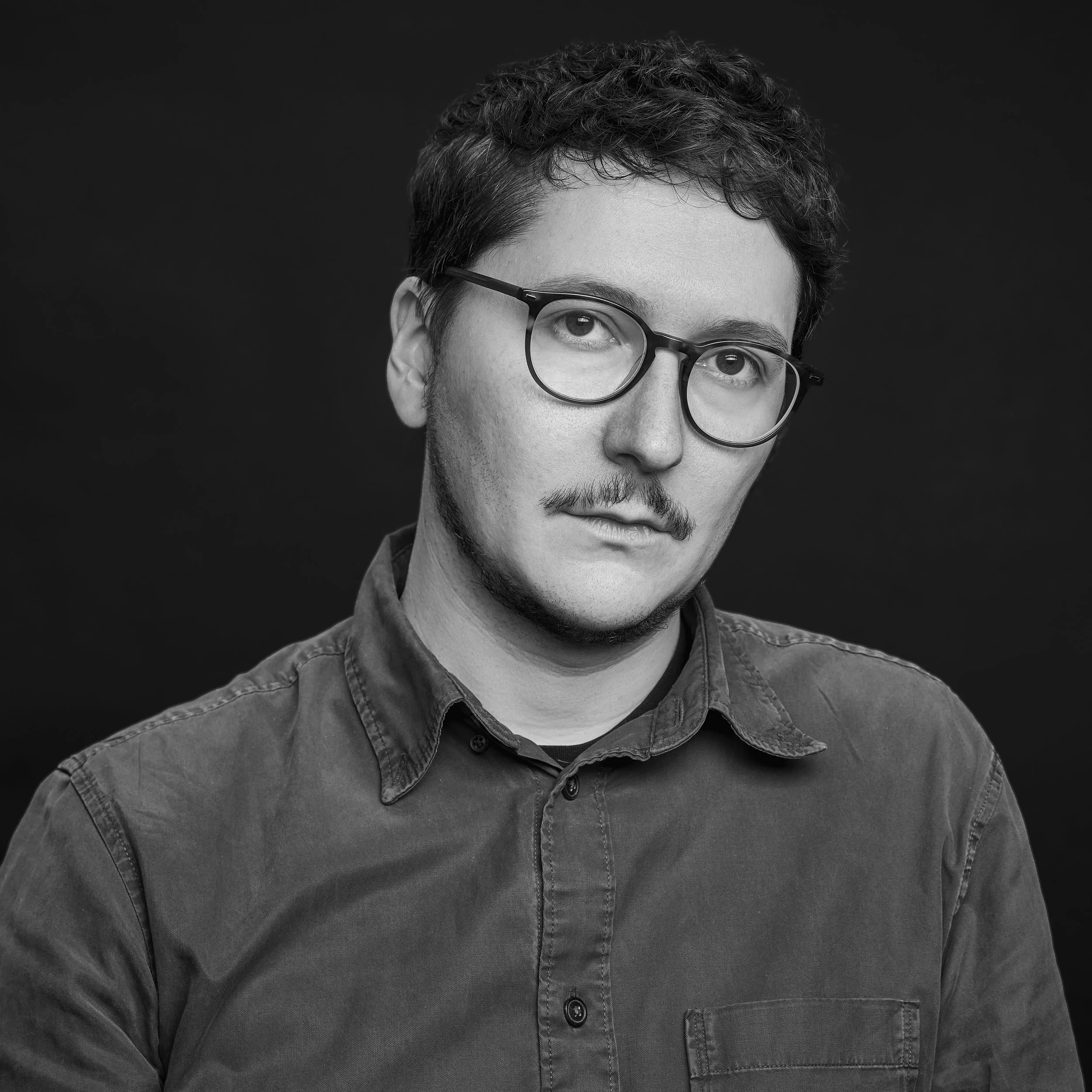 Portrait d'un homme jeune avec des cheveux bouclés, portant des lunettes, vêtu d'une chemise en jean, fond noir.