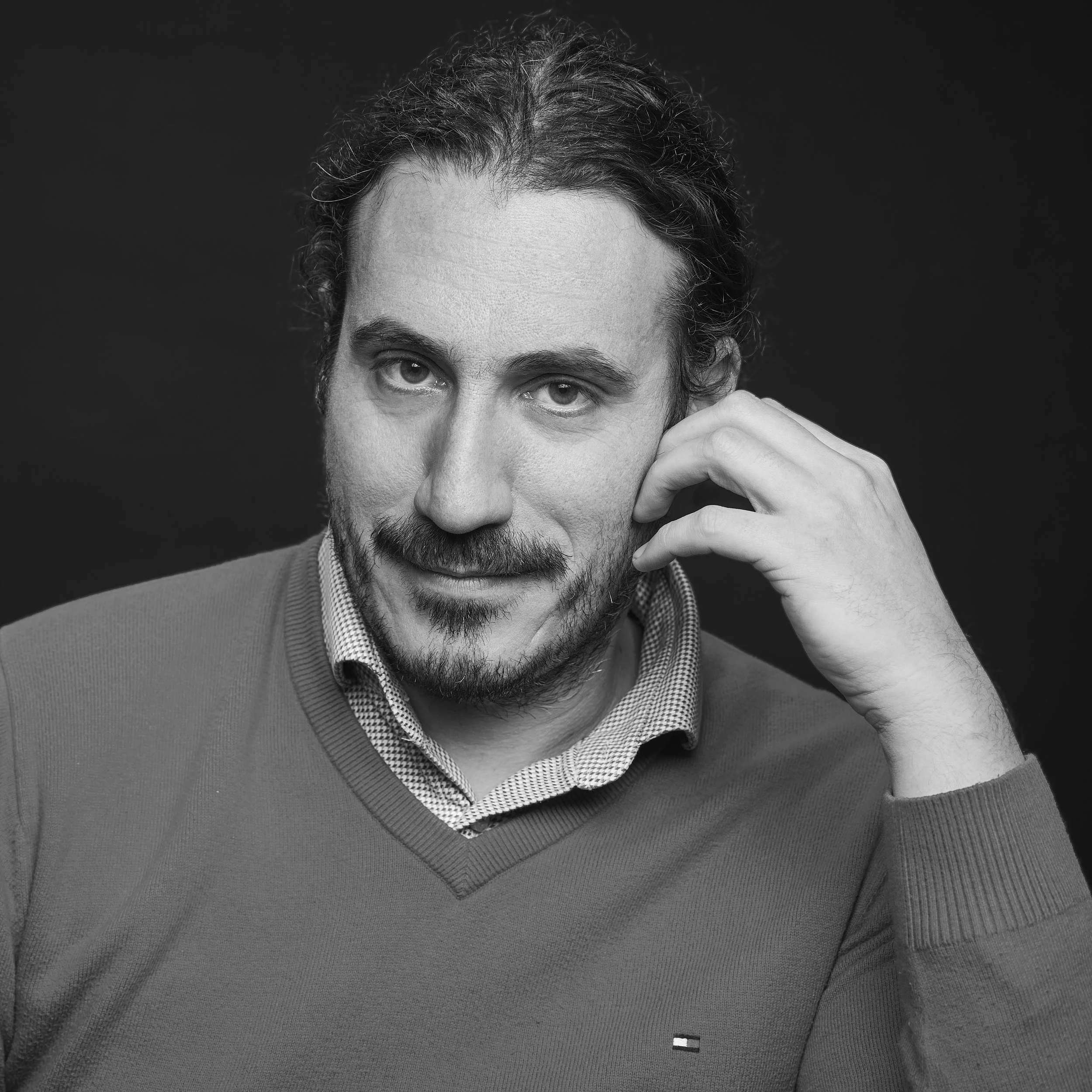Homme avec cheveux courts, barbe, portant un sweat gris et une chemise en dessous, un fond noir.