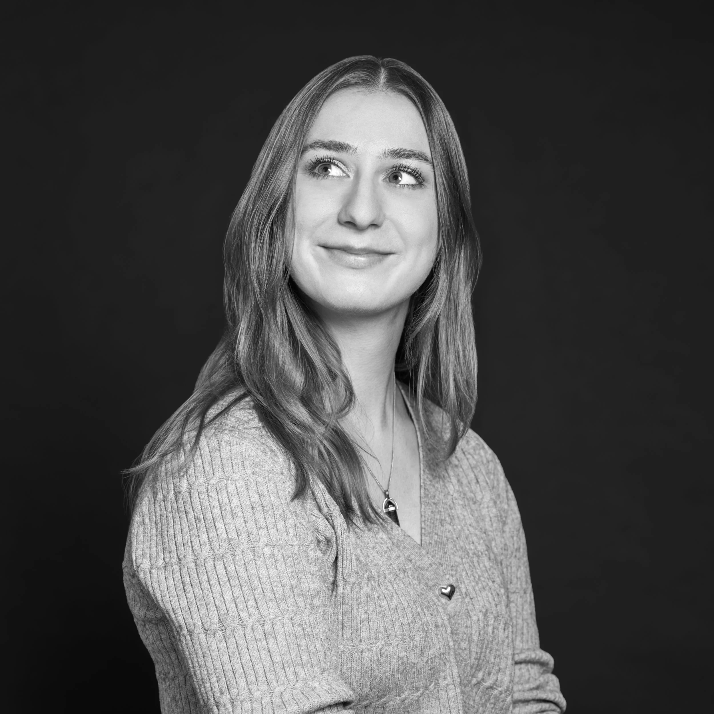 Portrait en noir et blanc d'une femme aux cheveux longs, portant un pull en maille et un collier, souriant légèrement, contre un fond sombre.