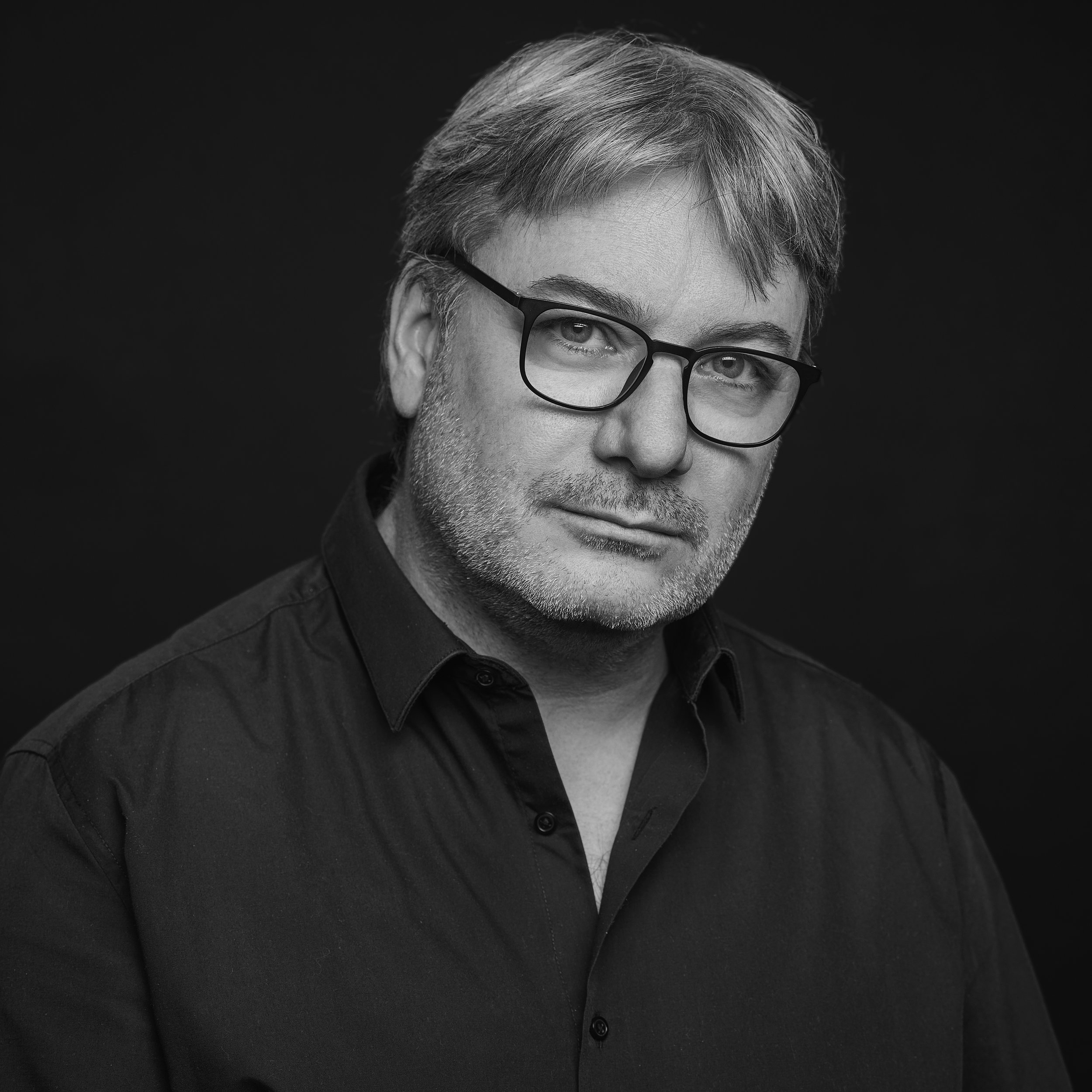 Portrait d'un homme portant des lunettes noires, barbe légère, cheveux gris, habillé d'une chemise noire, contre un fond noir.