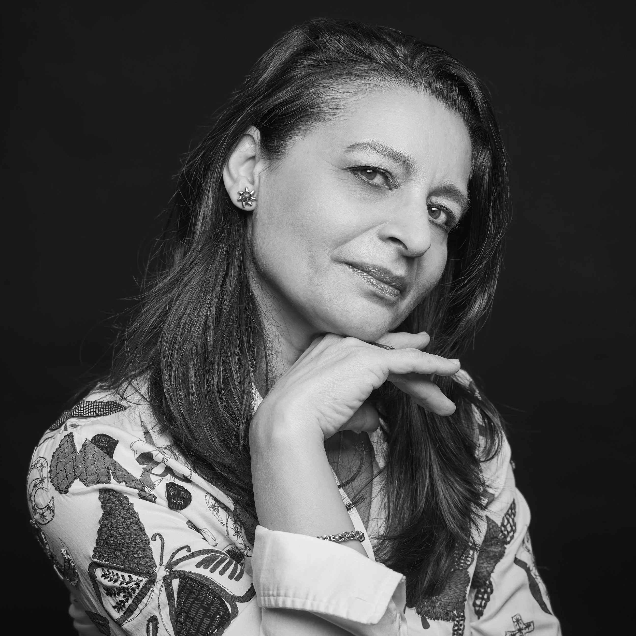 Portrait en noir et blanc d'une femme avec cheveux longs, portant des boucles d'oreilles et une chemise à motif de papillons. Elle pose sa main sous son menton, regardant légèrement vers la droite.